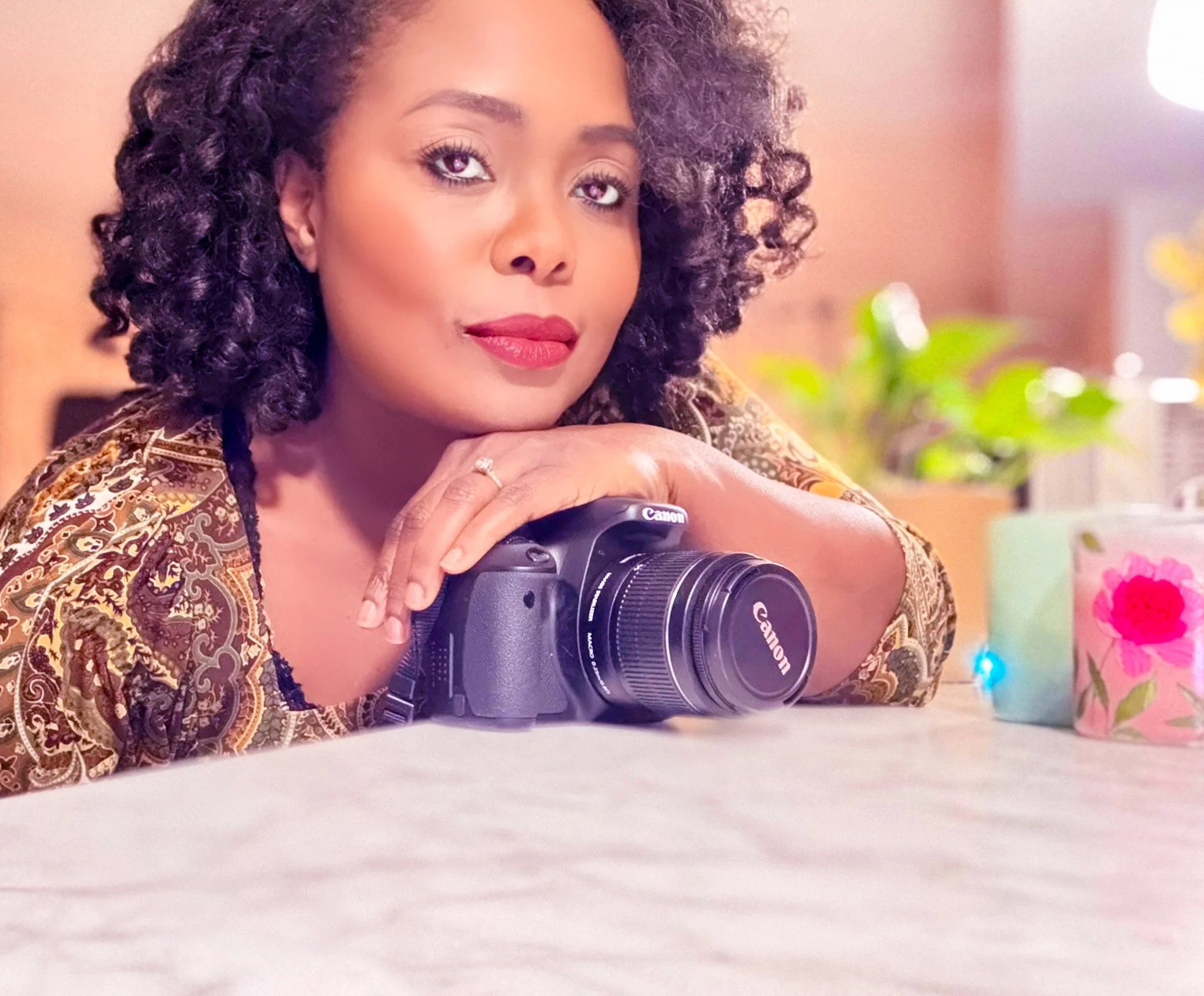 A woman with curly hair and red lipstick rests her chin on her hand, which is on a camera, on a table in an indoor setting with plants and colorful decor in the background.