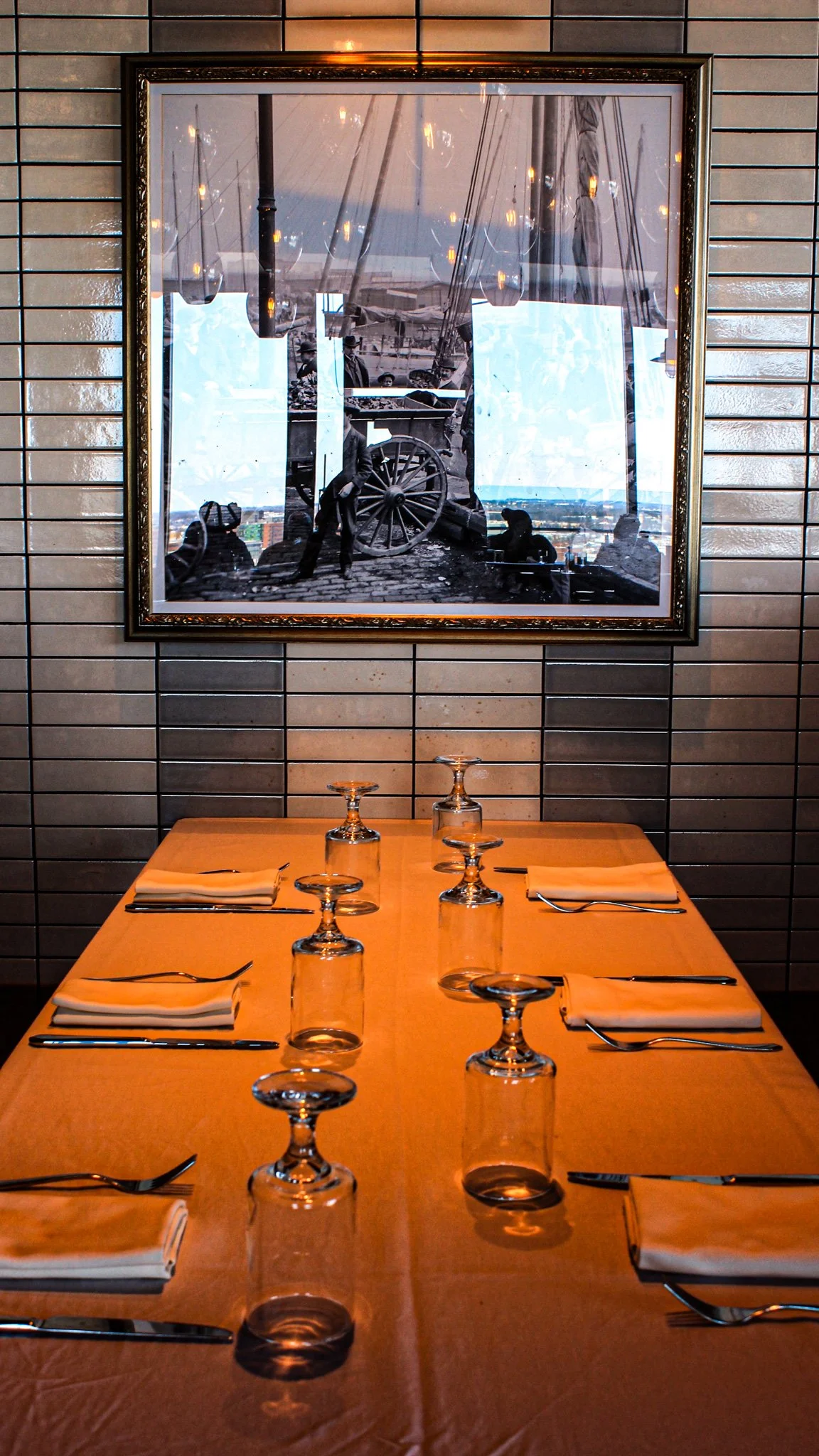 A restaurant table set with napkins, forks, knives, and upside-down glasses, with a large framed black and white photograph of a historical scene featuring a cannon and people, hanging on a tiled wall in the background.