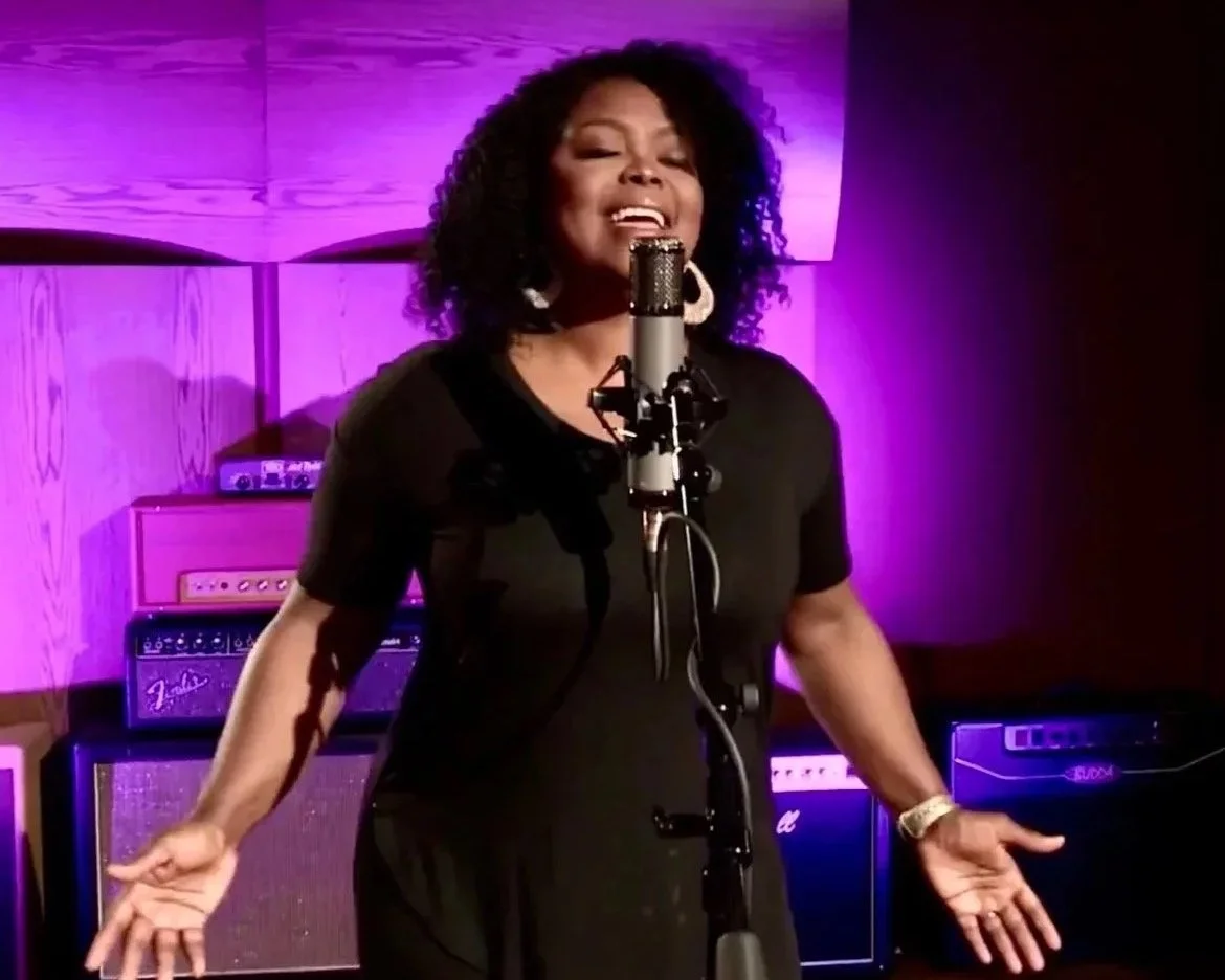 A woman singing into a microphone in a music studio with purple lighting, surrounded by guitar amplifiers.