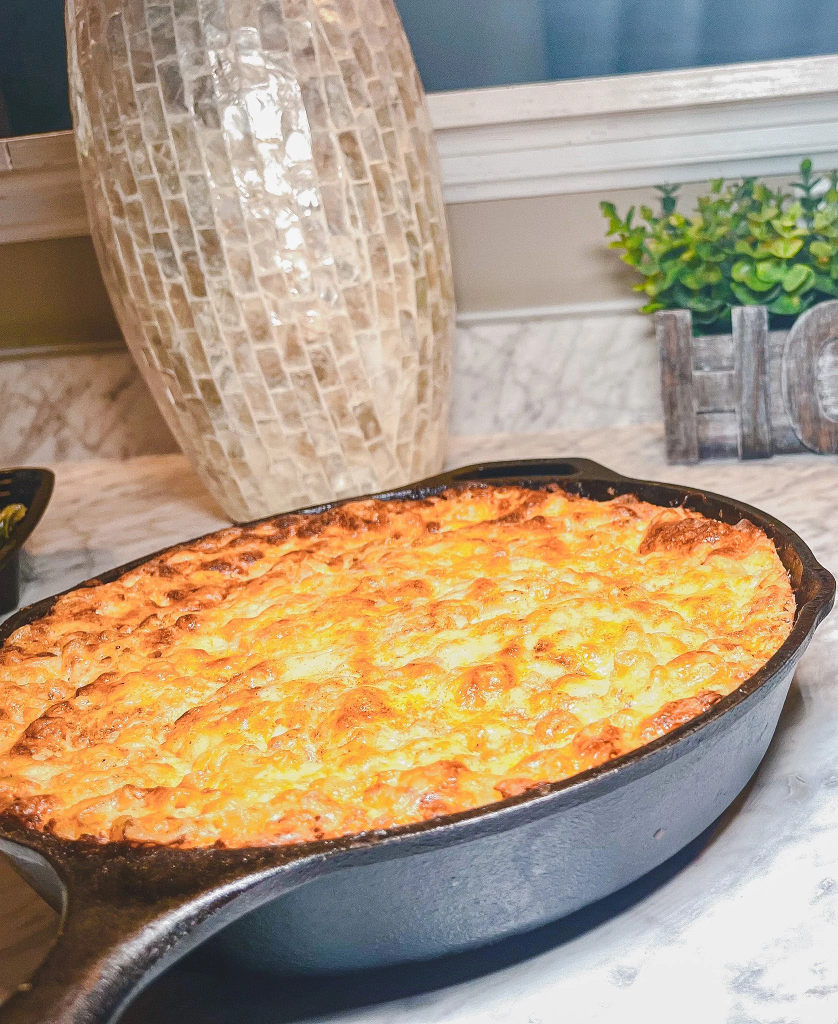 Baked cheesy dish in a black oval skillet on a marble countertop with decorative vases and plants in the background.