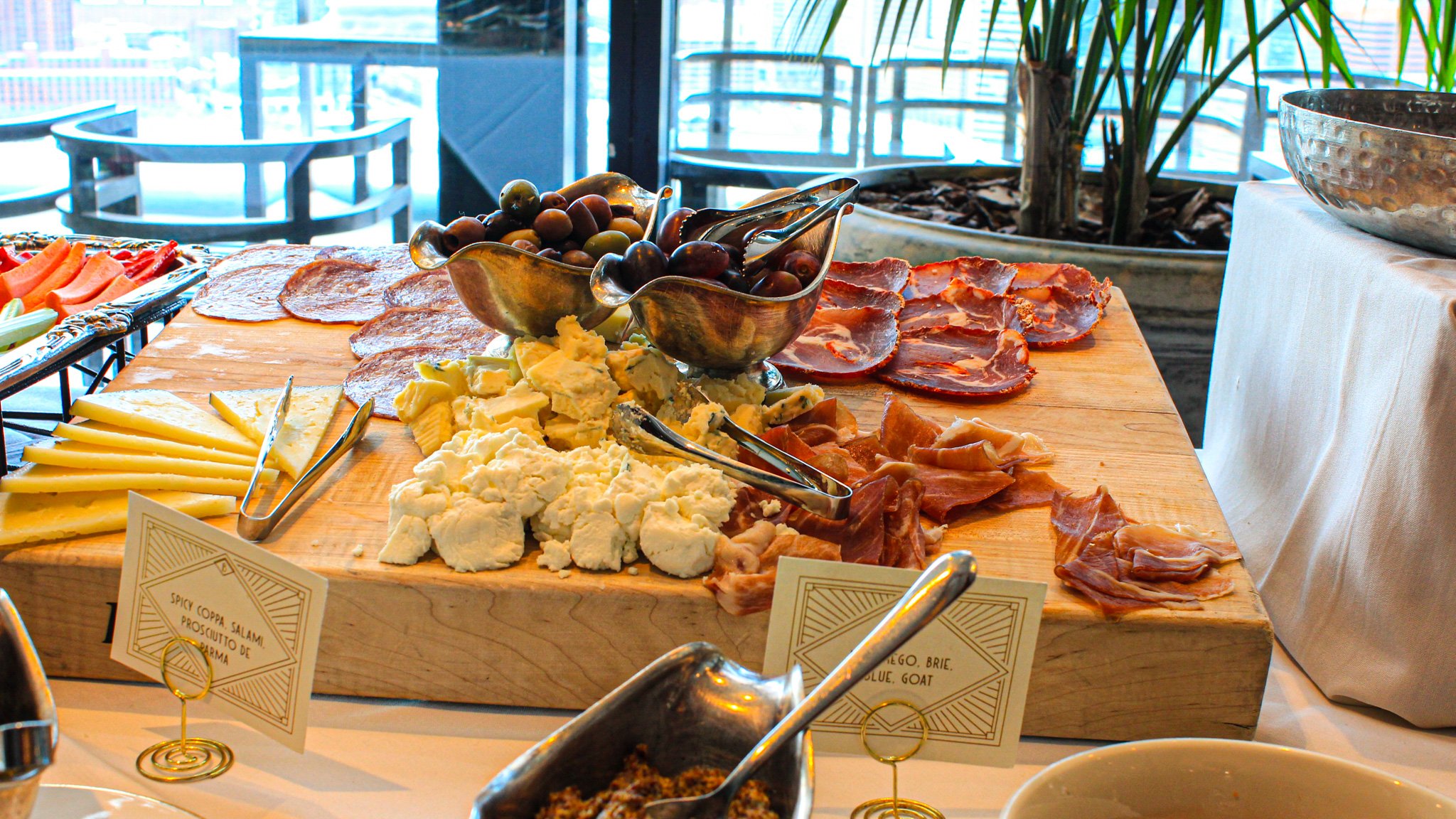 Wooden platter with an assortment of cheeses, cured meats, grapes, and other snacks at a buffet table.