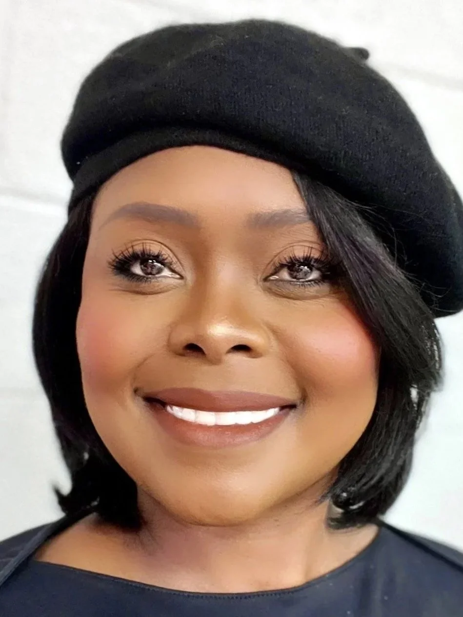 Close-up portrait of a woman wearing a black beret and a dark top, smiling with makeup and styled hair.