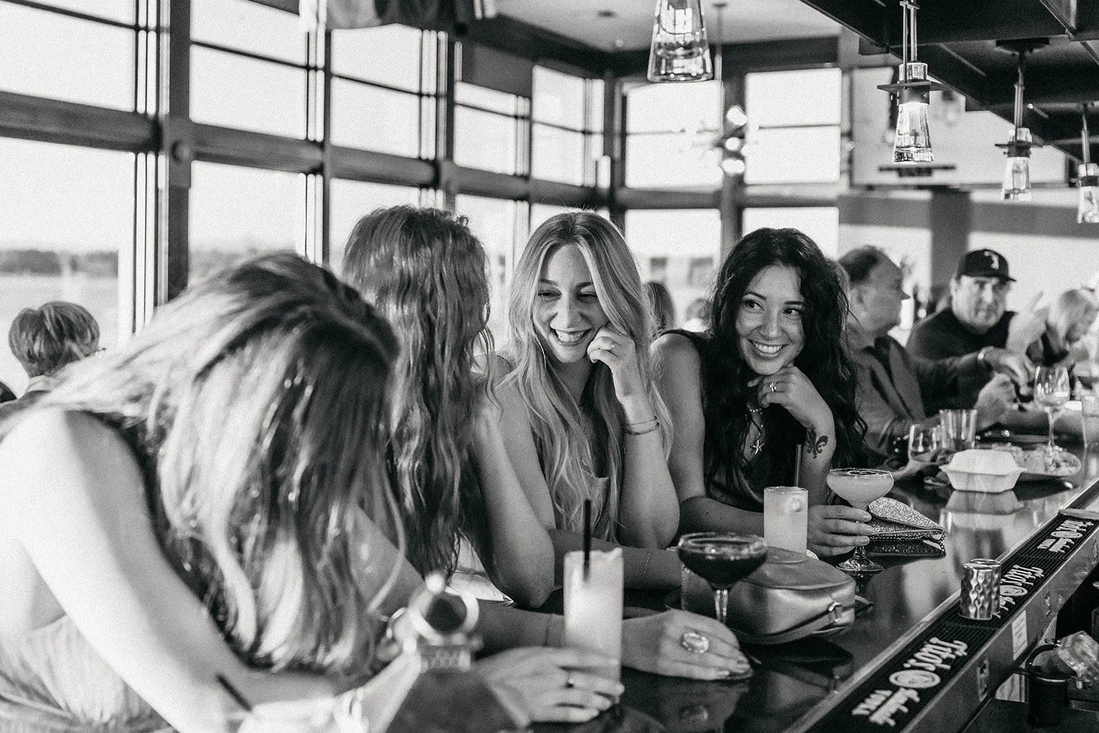 A group of women friends sitting at a bar, chatting and smiling, with drinks in front of them, in a modern restaurant with large windows.