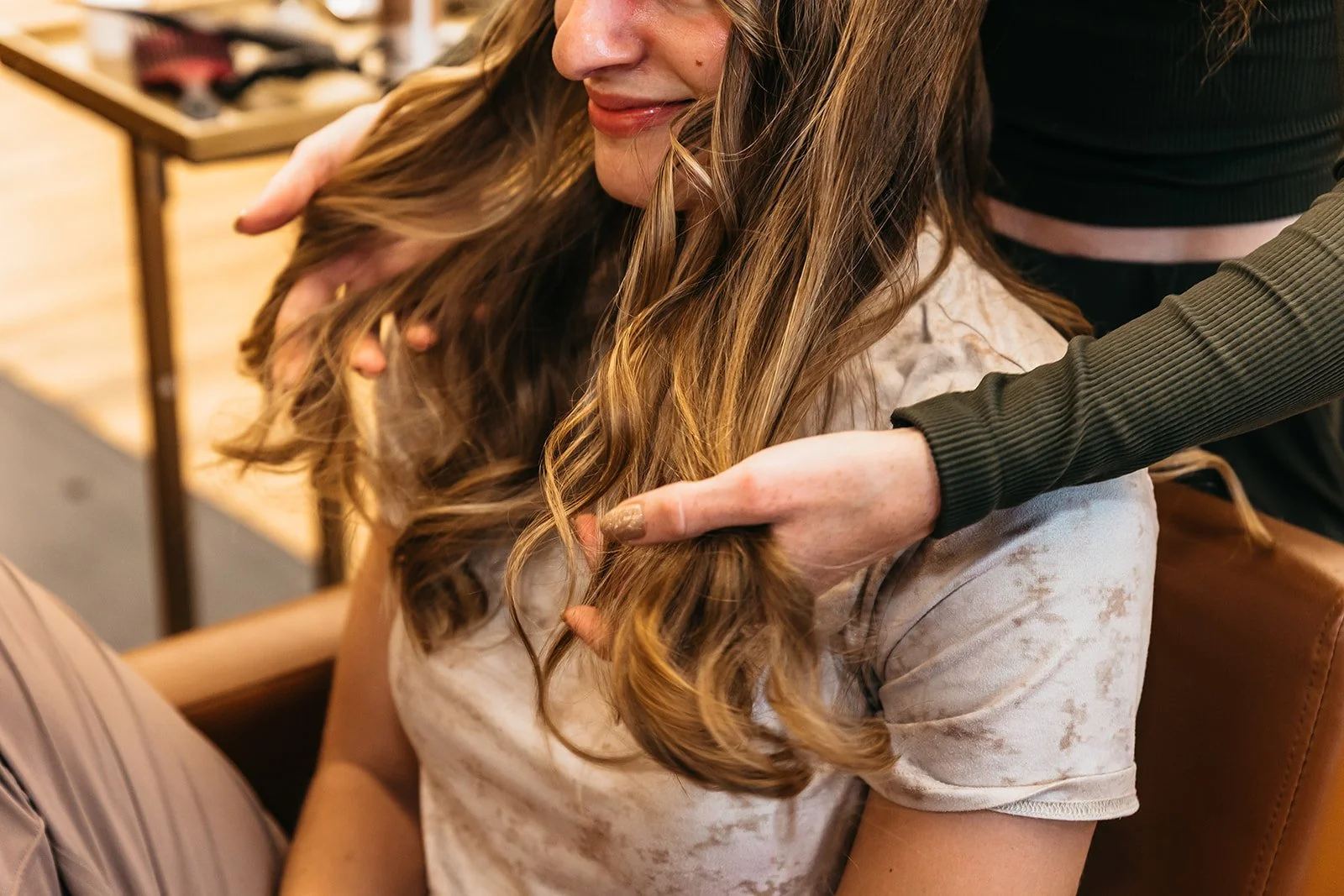 A woman with long, wavy, light brown hair sitting in a salon chair getting her hair styled by a stylist. The stylist's hands are visible, holding and working on her hair. The woman is smiling softly and wearing a light-colored, short-sleeved shirt.