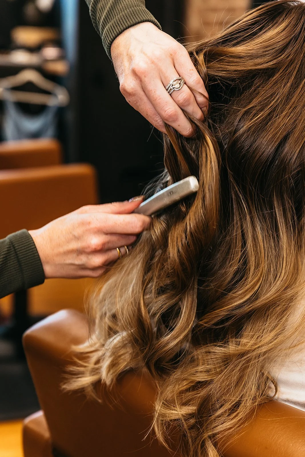 A hairstylist using a curling iron to style a woman's long, wavy, light brown hair in a salon.