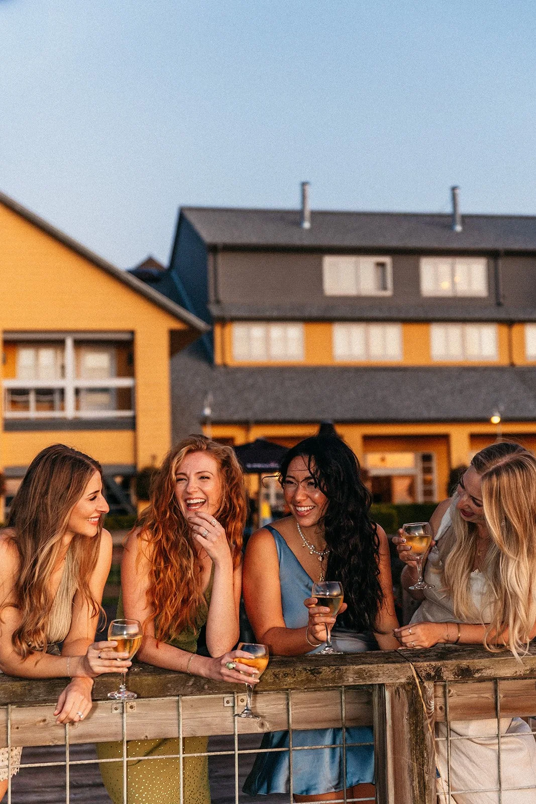 A group of five women enjoying drinks and laughing together on the PNW coast outdoors during sunset.
