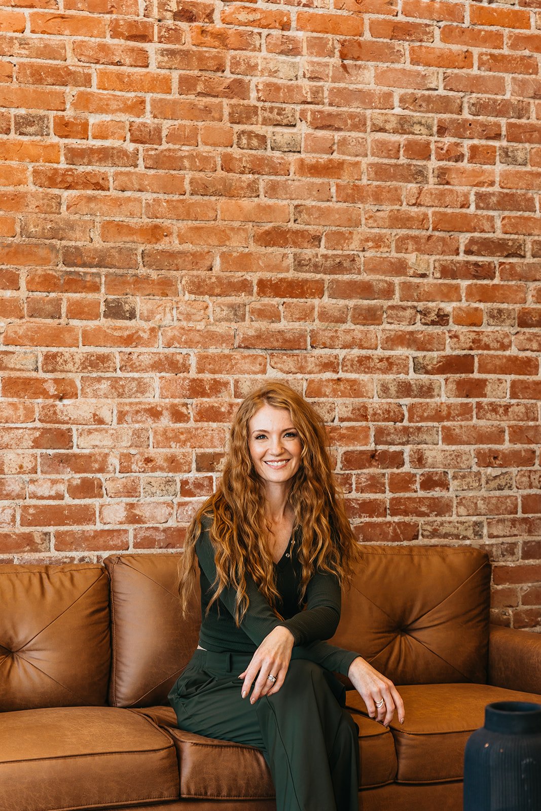 A woman with long, red, hair sitting on a brown leather couch in front of an exposed brick wall in downtown Lynden, WA, smiling at the camera.