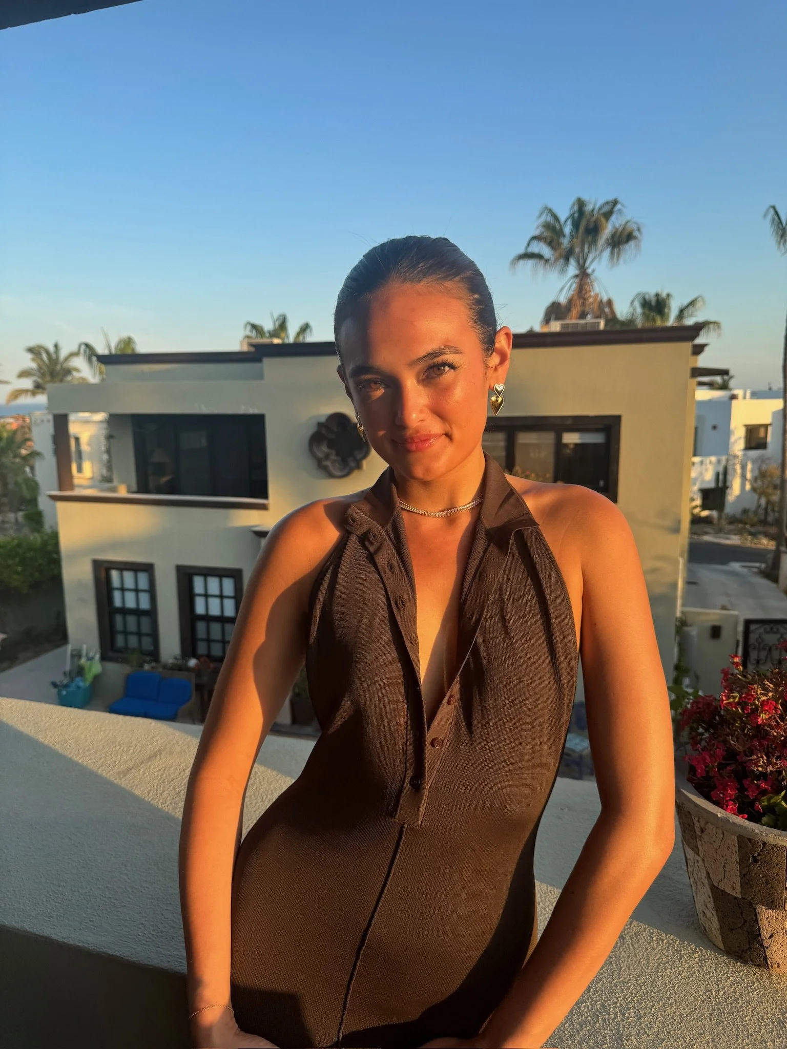 A woman with dark hair tied back, wearing a sleeveless, partially unbuttoned brown dress, stands on a balcony during sunset, with a modern house and palm trees in the background.