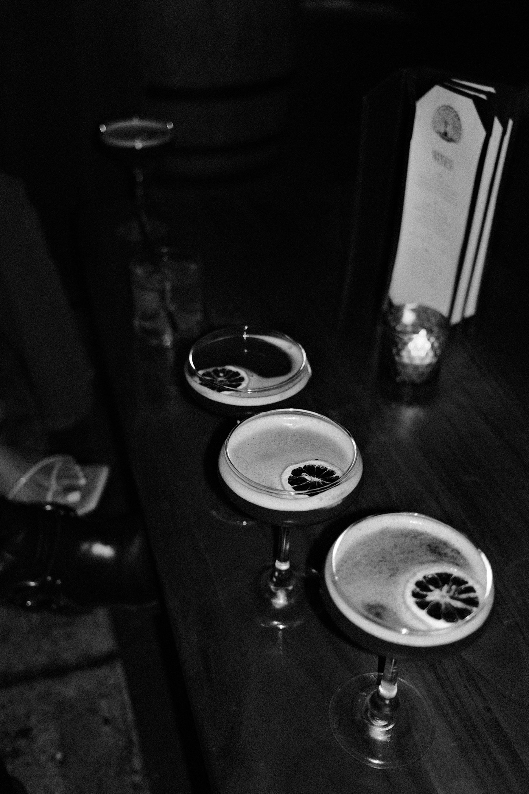 Three cocktails with lemon slices garnishes arranged in a line on a dark table in a dimly lit bar setting.
