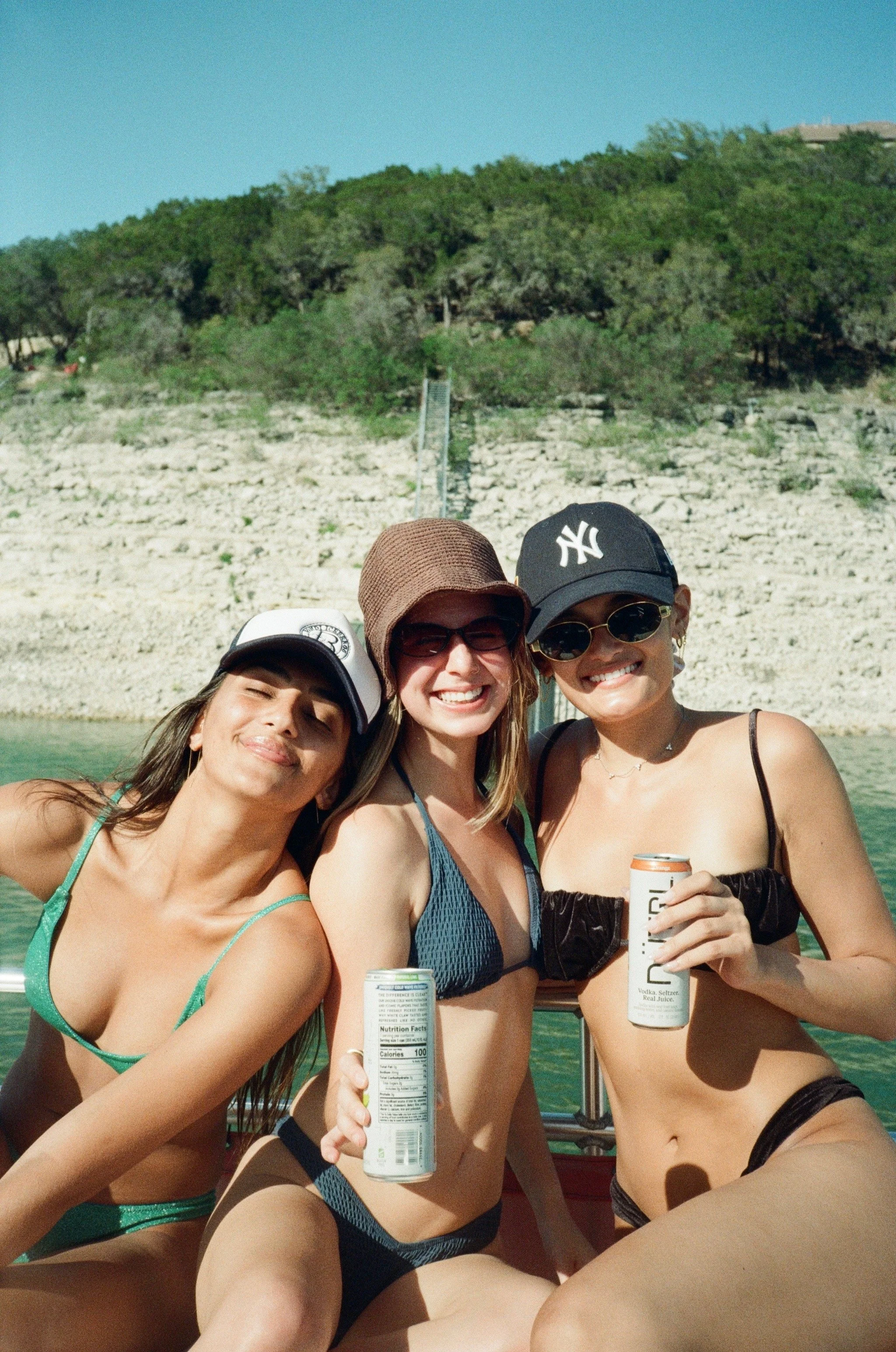 Three smiling women in swimsuits and hats sitting on a boat, holding canned drinks, with a rocky and green hillside in the background.