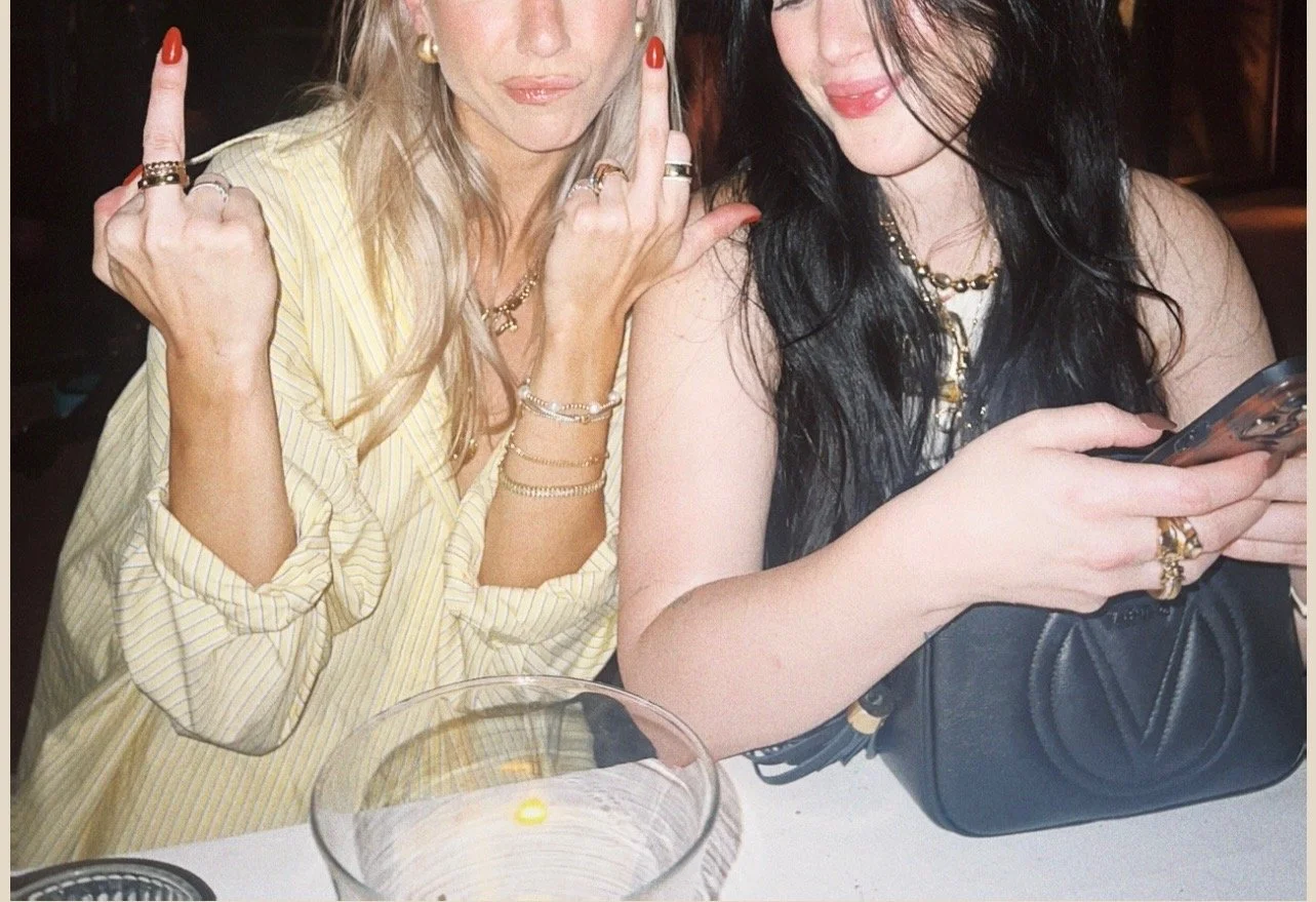 Two women sitting at a table. One woman has blonde hair and is making a peace sign with her right hand, some rings on her fingers. The other woman has black hair and is looking at her phone, with a black purse on the table in front of her.