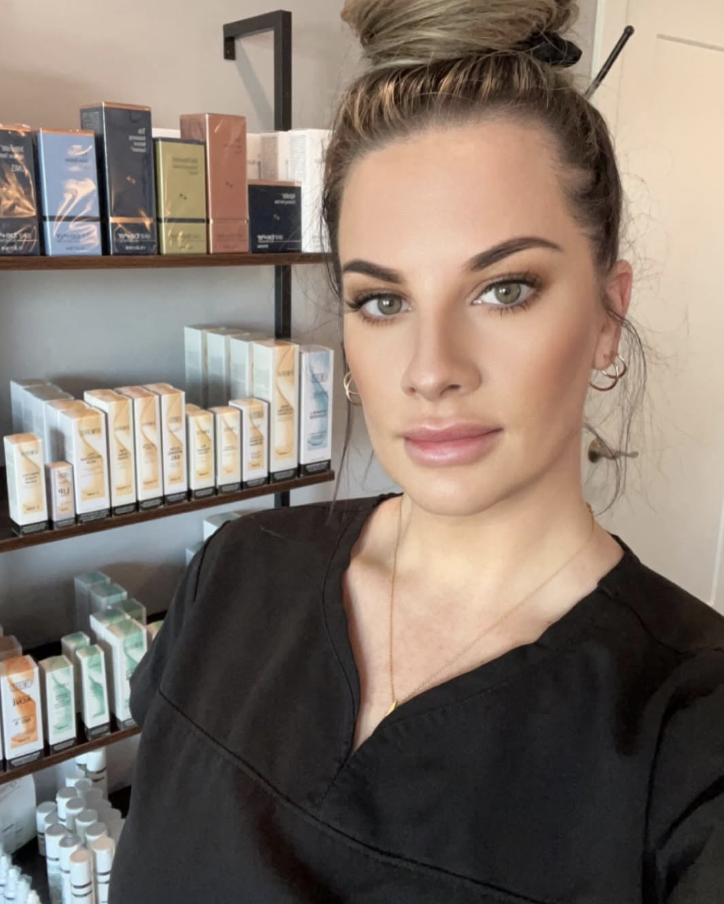 A woman with light skin and green eyes taking a selfie in front of shelves stocked with skincare or cosmetic products.
