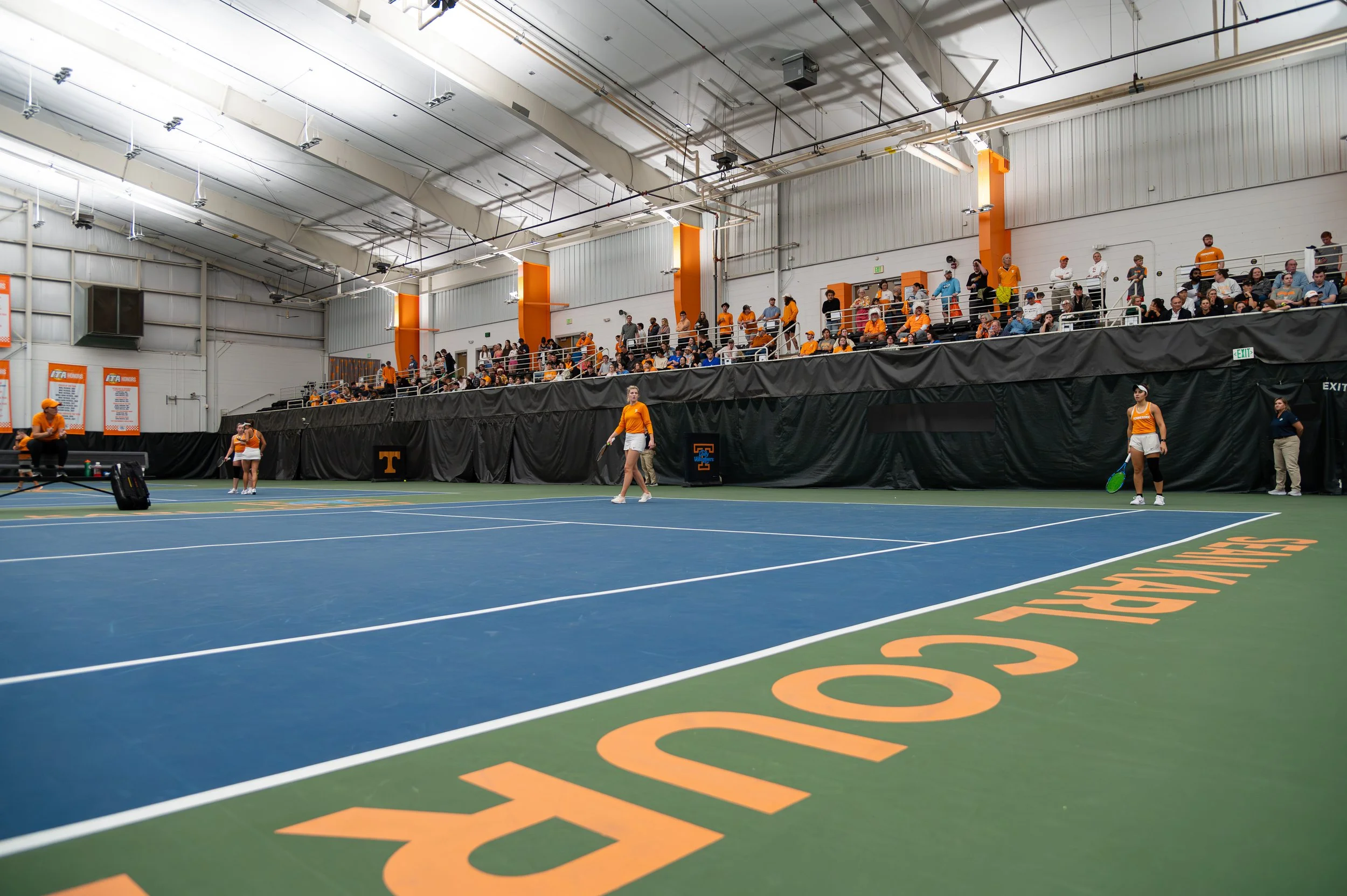 An indoor tennis court with players and spectators, featuring orange and white color theme representing the University of Tennessee.