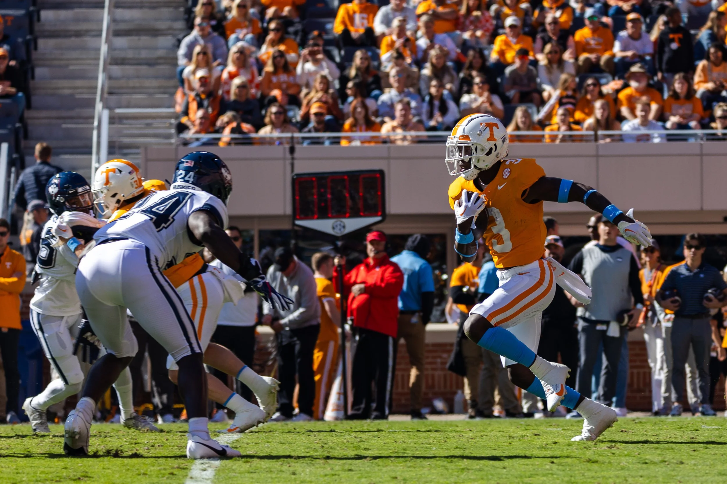 An American football game with a player in orange jersey running with the ball while other players in white jerseys defend. The crowd in the stands is dressed mostly in orange and white.