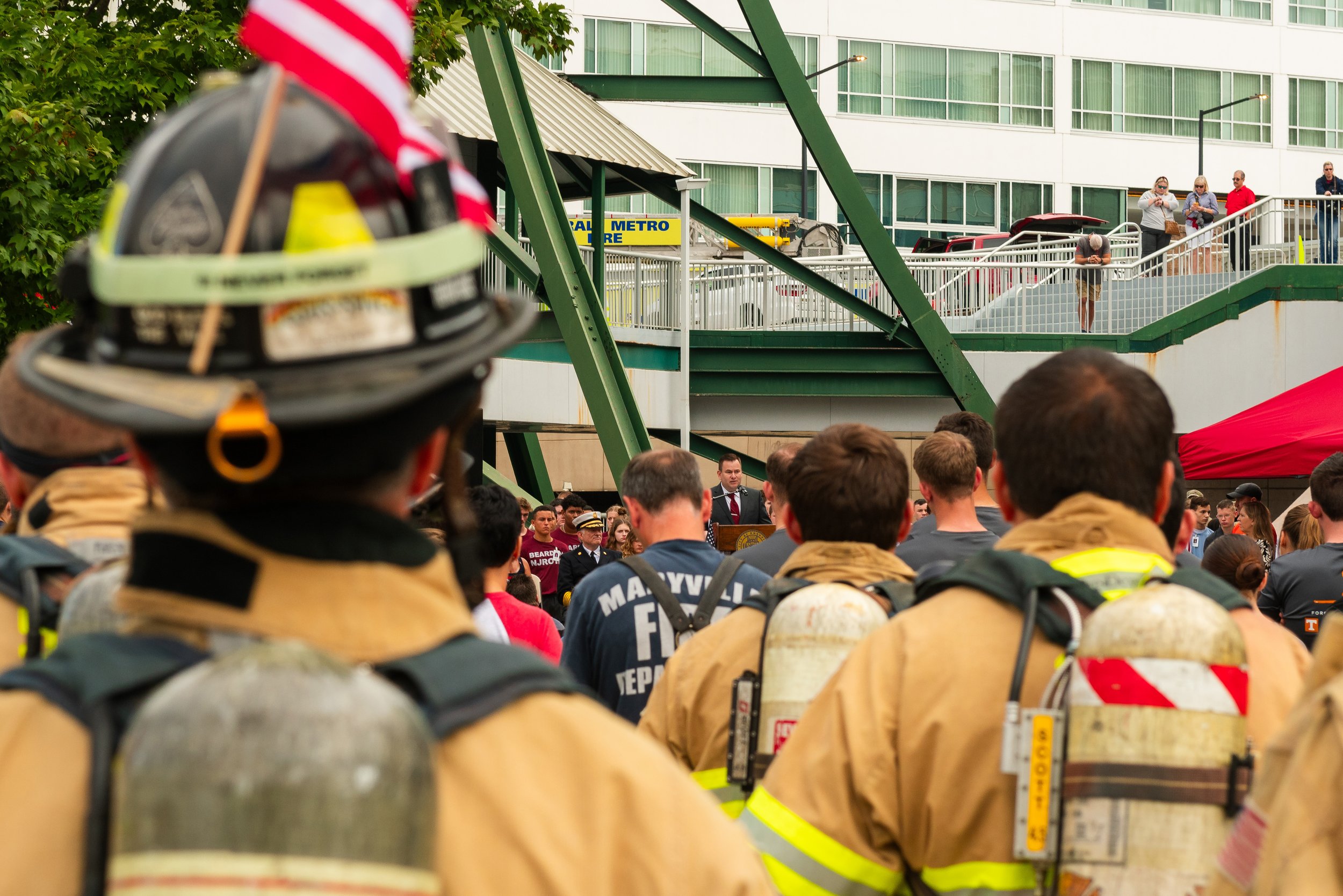 Firefighters in uniform attending a public event, with a speaker addressing the crowd and people standing on a raised platform in the background.