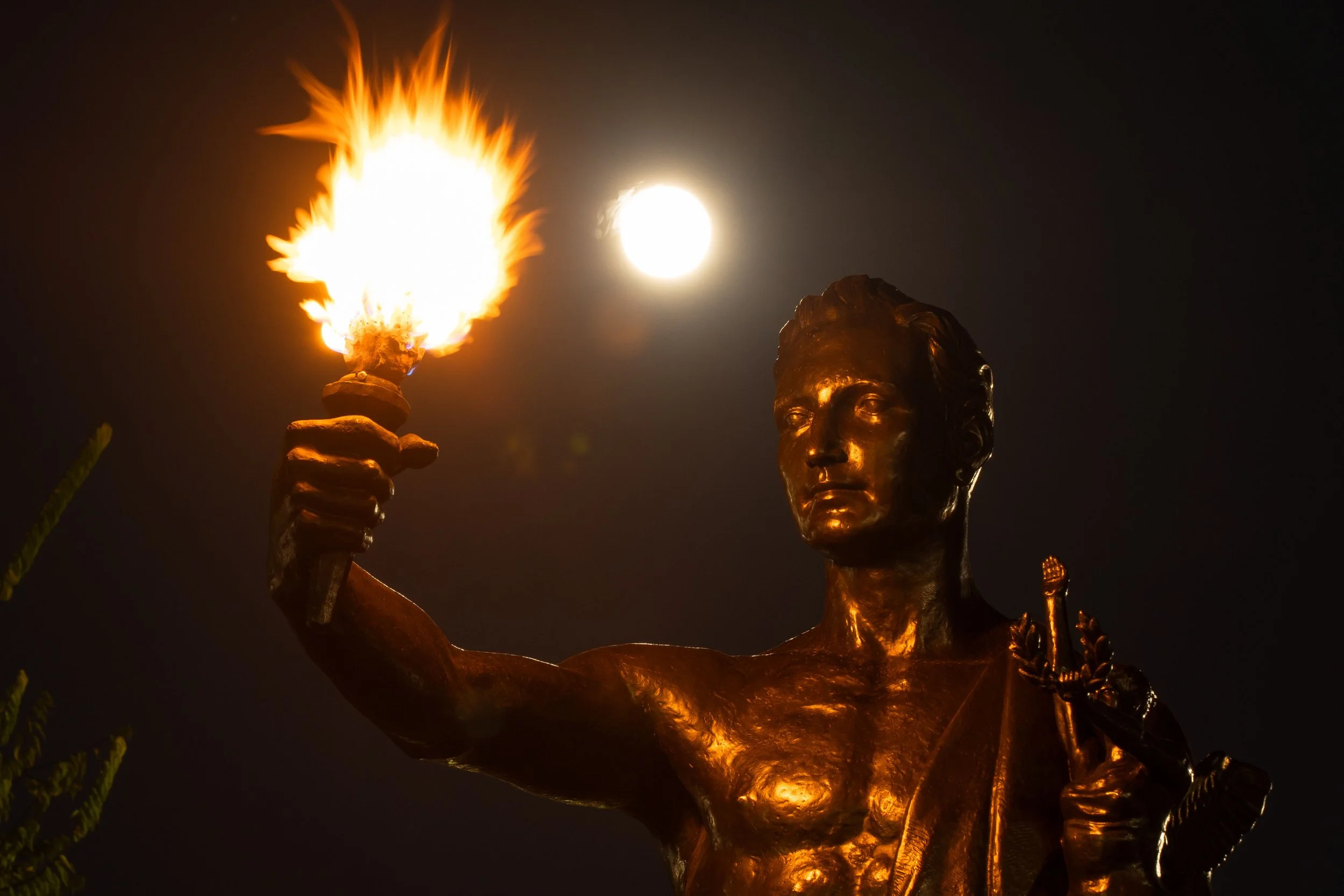 Bronze statue of a person holding a torch with fire, illuminated by moonlight at night