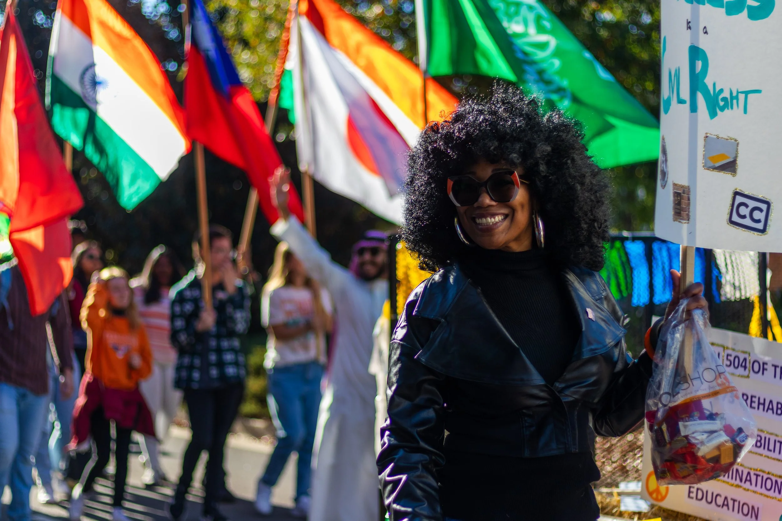 A woman with curly black hair, sunglasses, and hoop earrings smiling at a parade. She is wearing a black leather jacket and holding a plastic bag filled with various items. Behind her, people are holding flags of different countries and a colorful pa