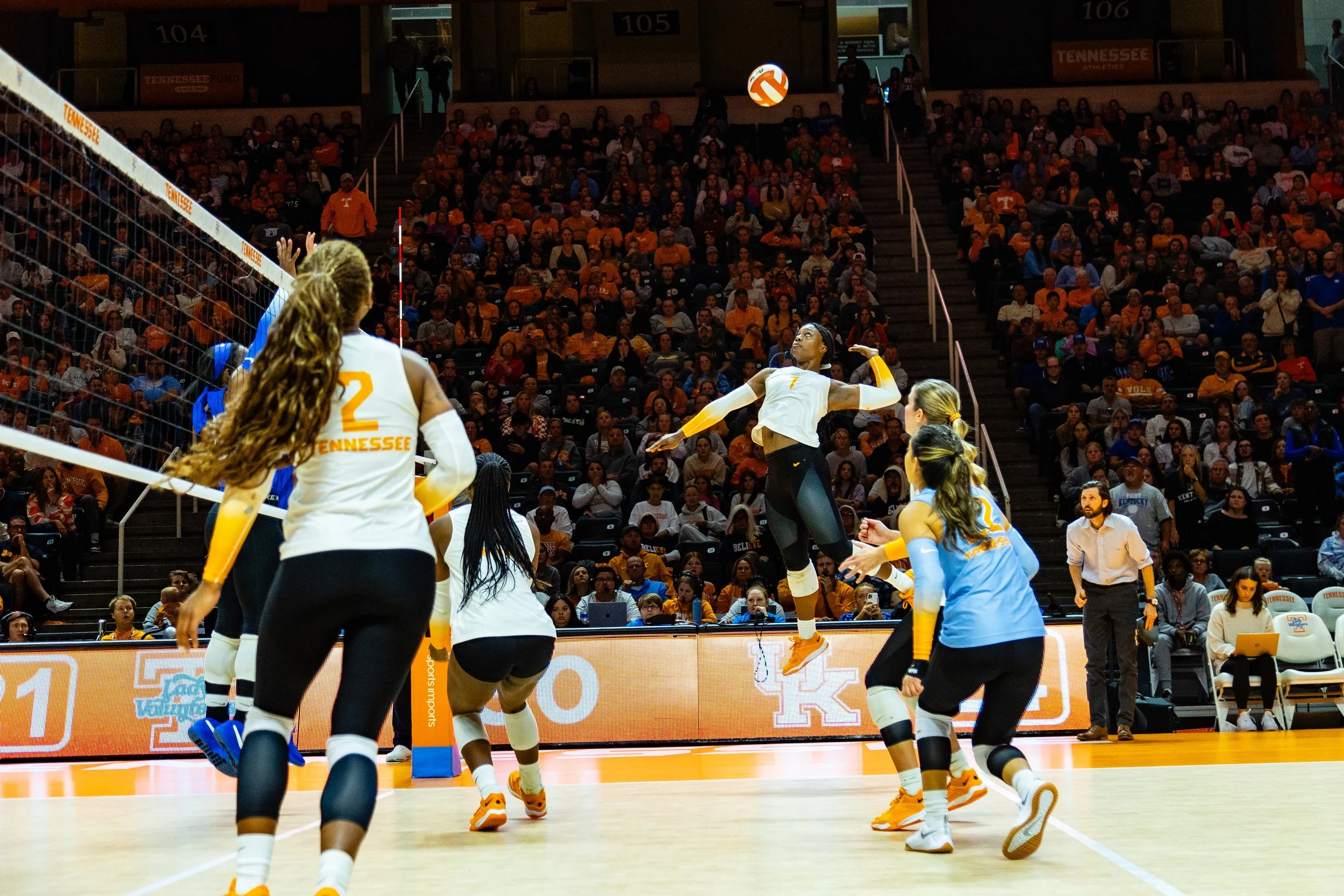 Tennessee women's volleyball team during a game, with one player jumping to hit the ball over the net, and other players preparing to receive or block, in an indoor stadium filled with spectators.