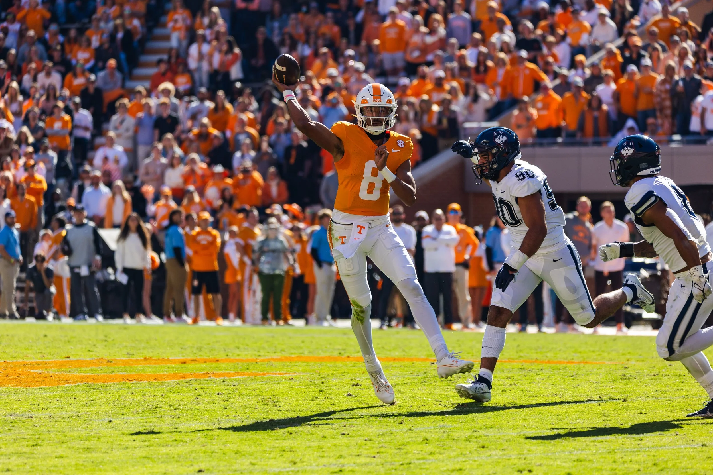 A college football game with a Tennessee player in orange uniform diving to catch a football, while a Michigan player in white uniform is running towards him, and a teammate nearby, on a green field with a crowd in orange and white in the background.