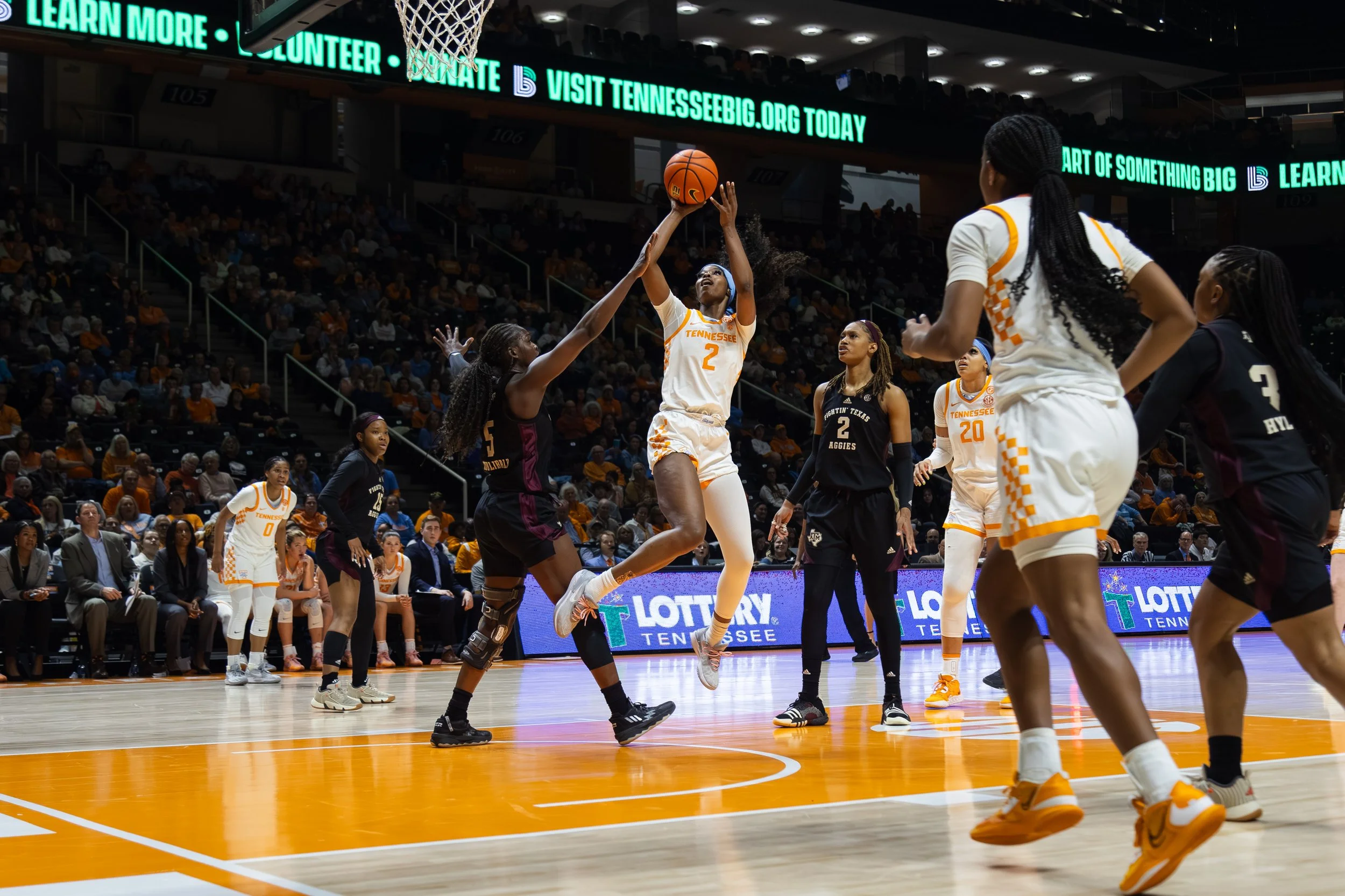 Group of female basketball players in action during a game, with one player jumping to shoot the ball, on a basketball court with spectators in the background.