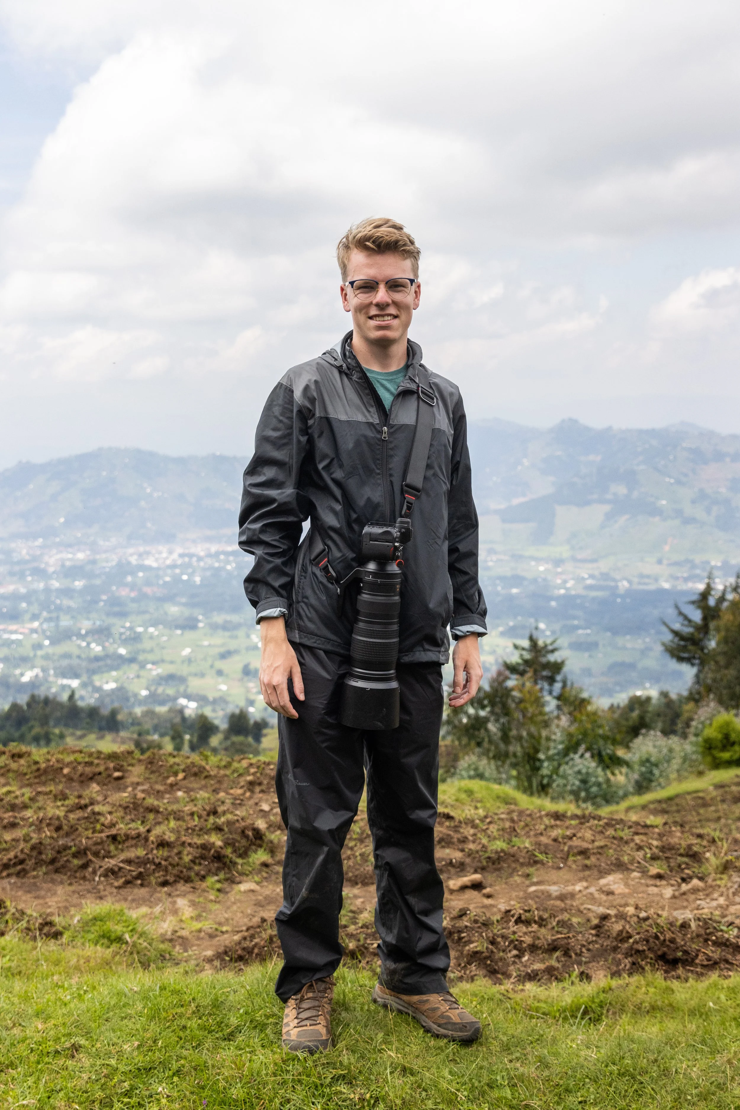 A young man stands outdoors on a grassy hilltop during daytime, dressed in outdoor gear with a large camera hanging from his neck, scenic landscape with rolling hills and cloudy sky in the background.