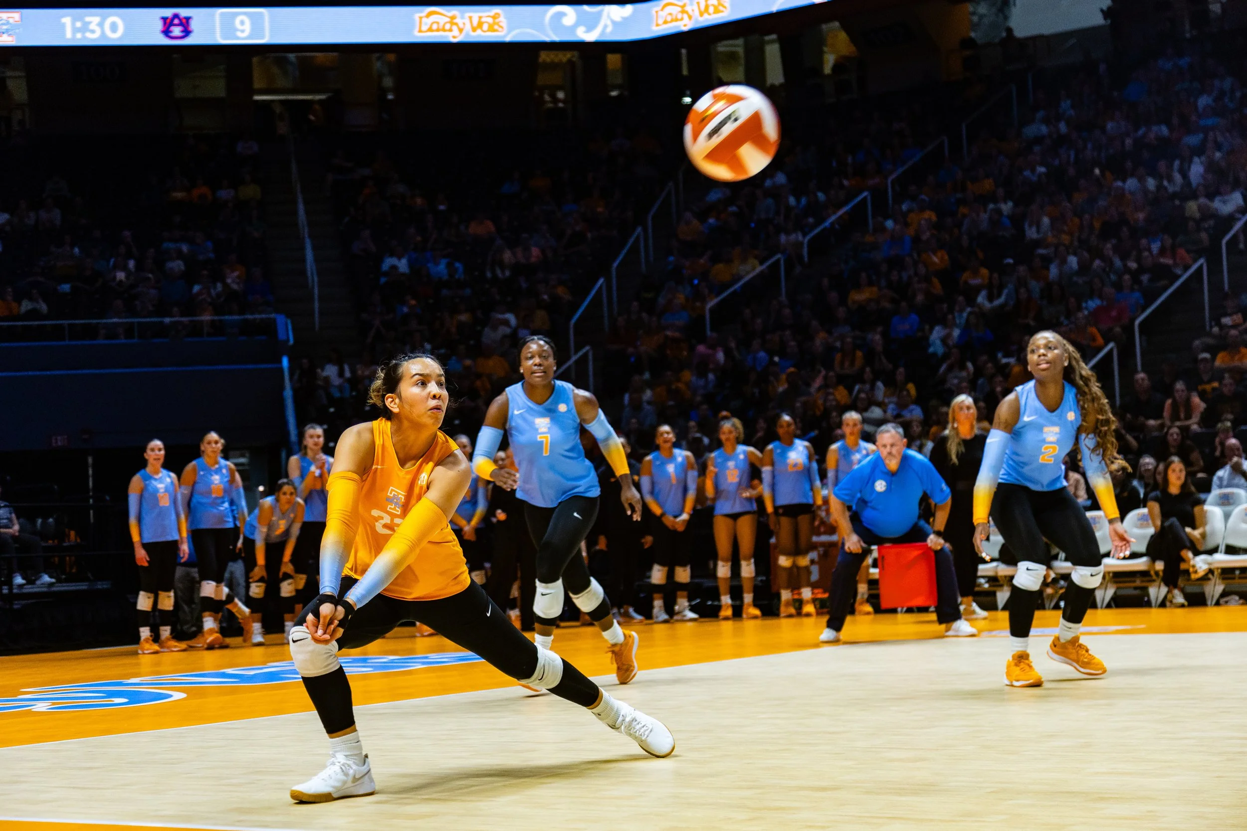 Women's volleyball game in progress, players in yellow and blue uniforms on the court, audience in the background.