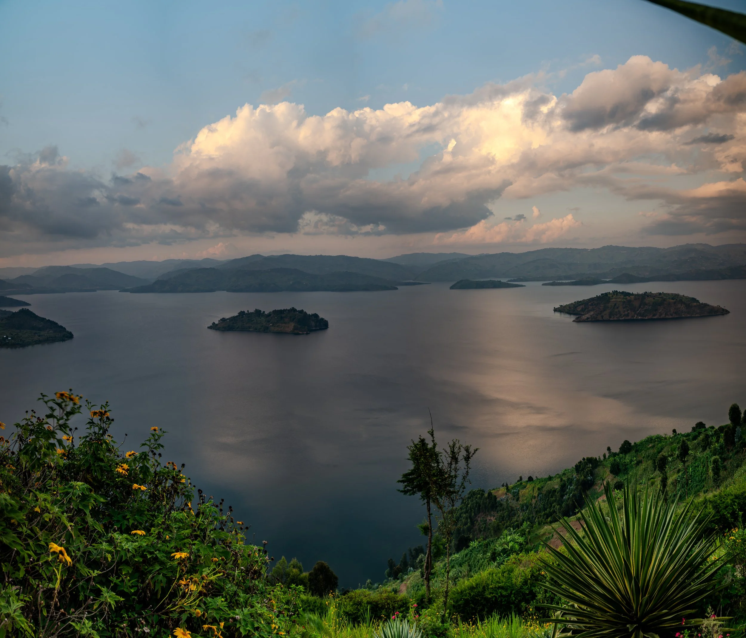 Scenic view of a lake with small islands, surrounded by lush green hills and a cloudy sky at sunset.