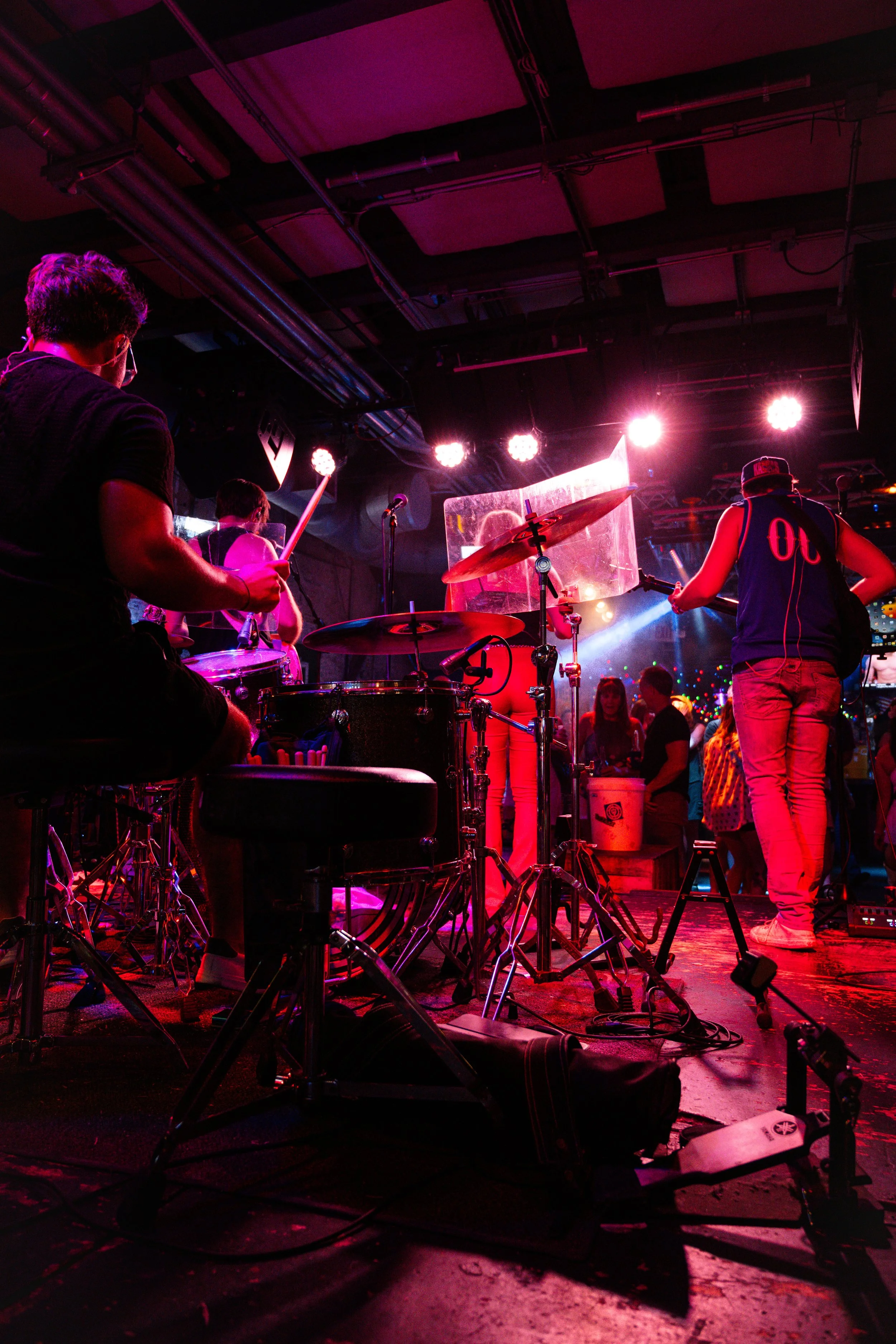 Band performing on stage with drums and microphones under colorful stage lights, with audience members in the background.