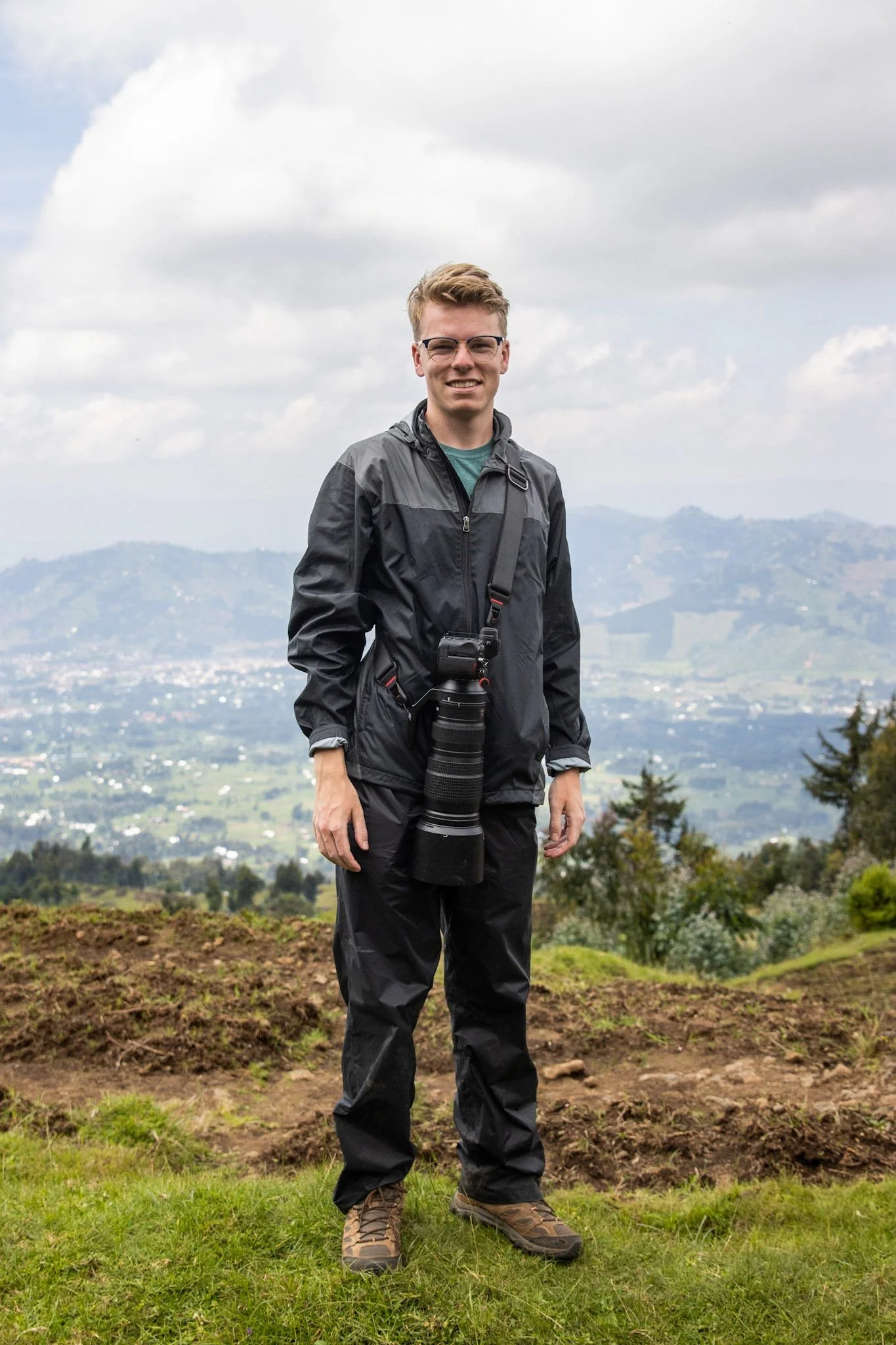 A man standing outdoors on a grassy hilltop with a camera hanging from his neck, wearing a black jacket and pants, with a panoramic landscape of hills and a cloudy sky in the background.