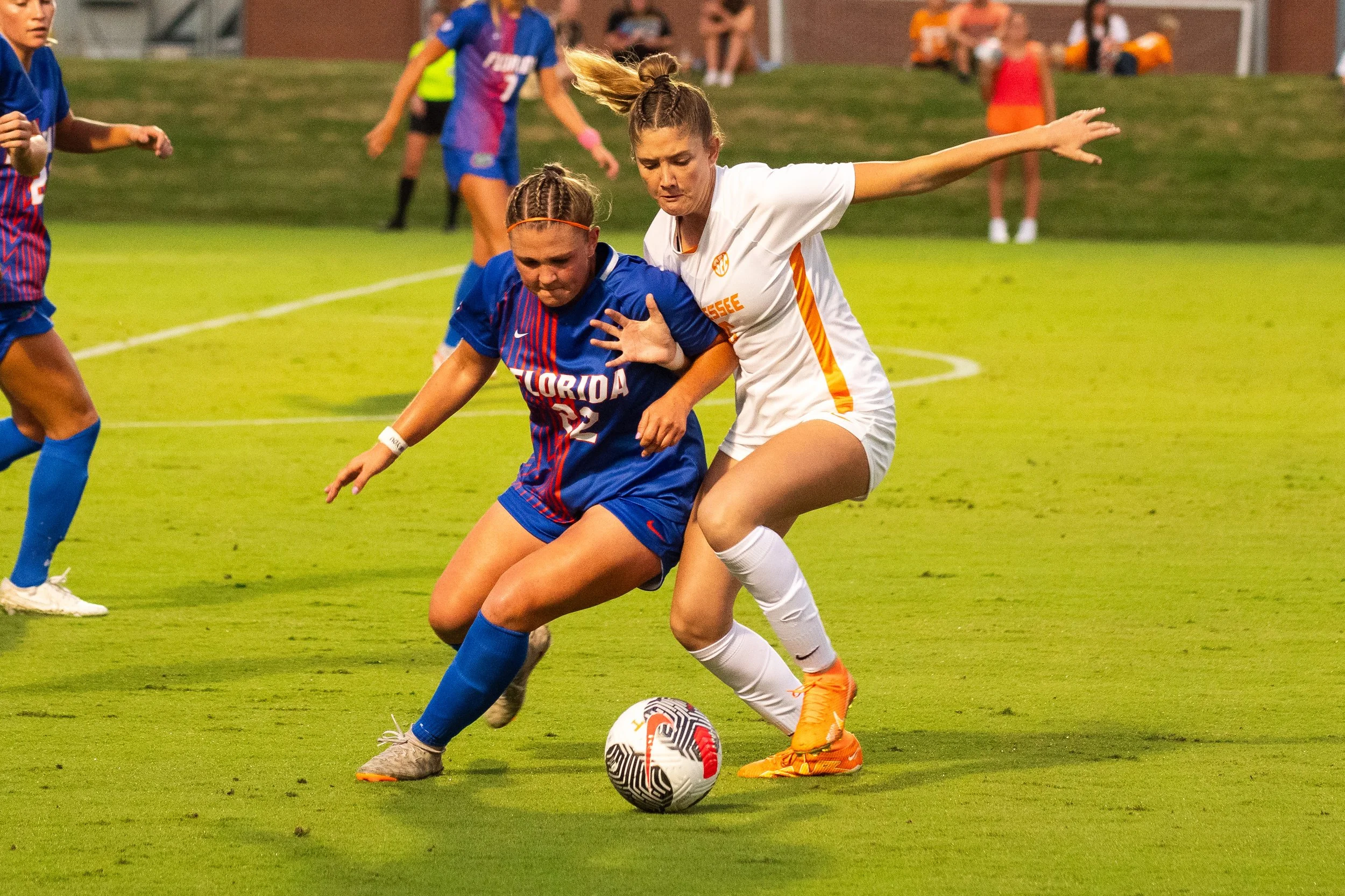 Two female soccer players competing for the ball during a game, with other players and spectators in background.