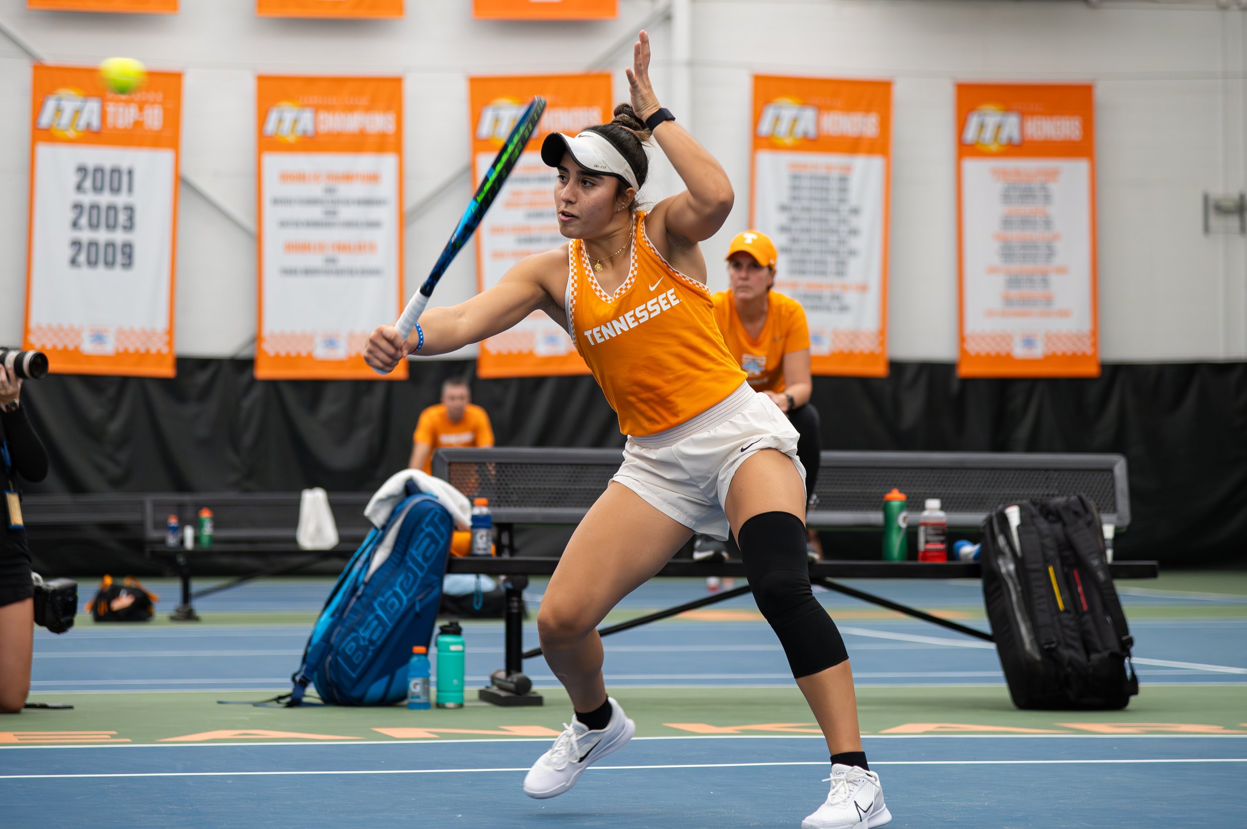 A female tennis player from Tennessee in orange attire preparing to hit a tennis ball on an indoor court.