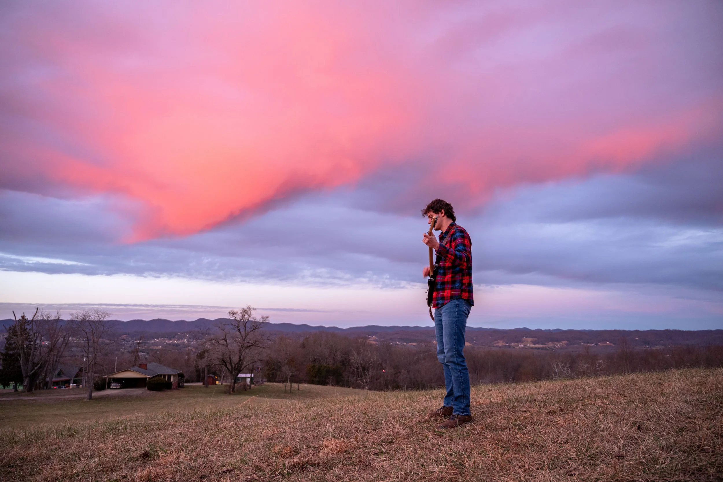 A man in a red and blue plaid shirt and jeans playing electric guitar outdoors during sunset, with pink and purple clouds overhead and a rural landscape with trees and houses in the background.