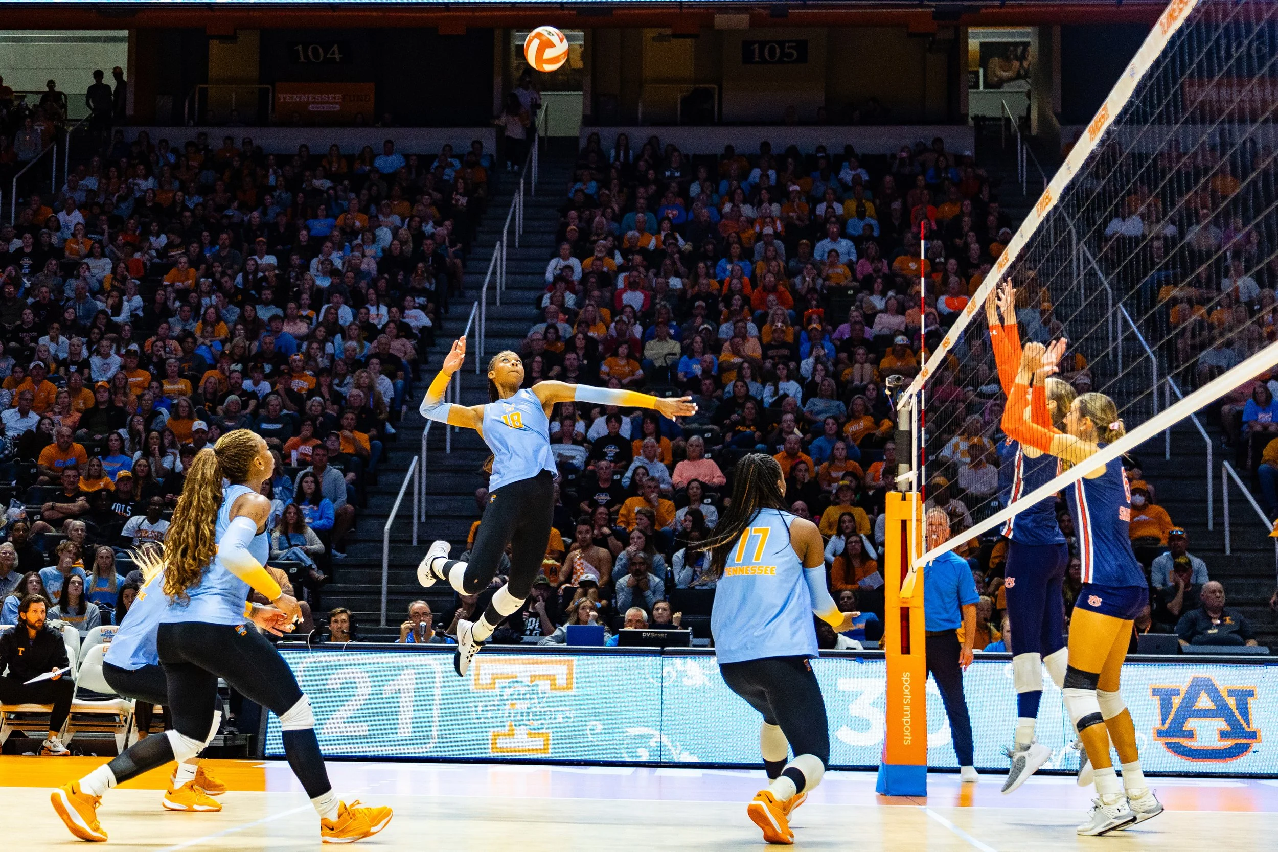A women's volleyball game in progress at an indoor stadium with spectators in the background. A player in a light blue jersey, number 18, is jumping to hit the ball. Other players in light blue jerseys and two players in dark blue and orange jerseys are near the net, preparing for the play.