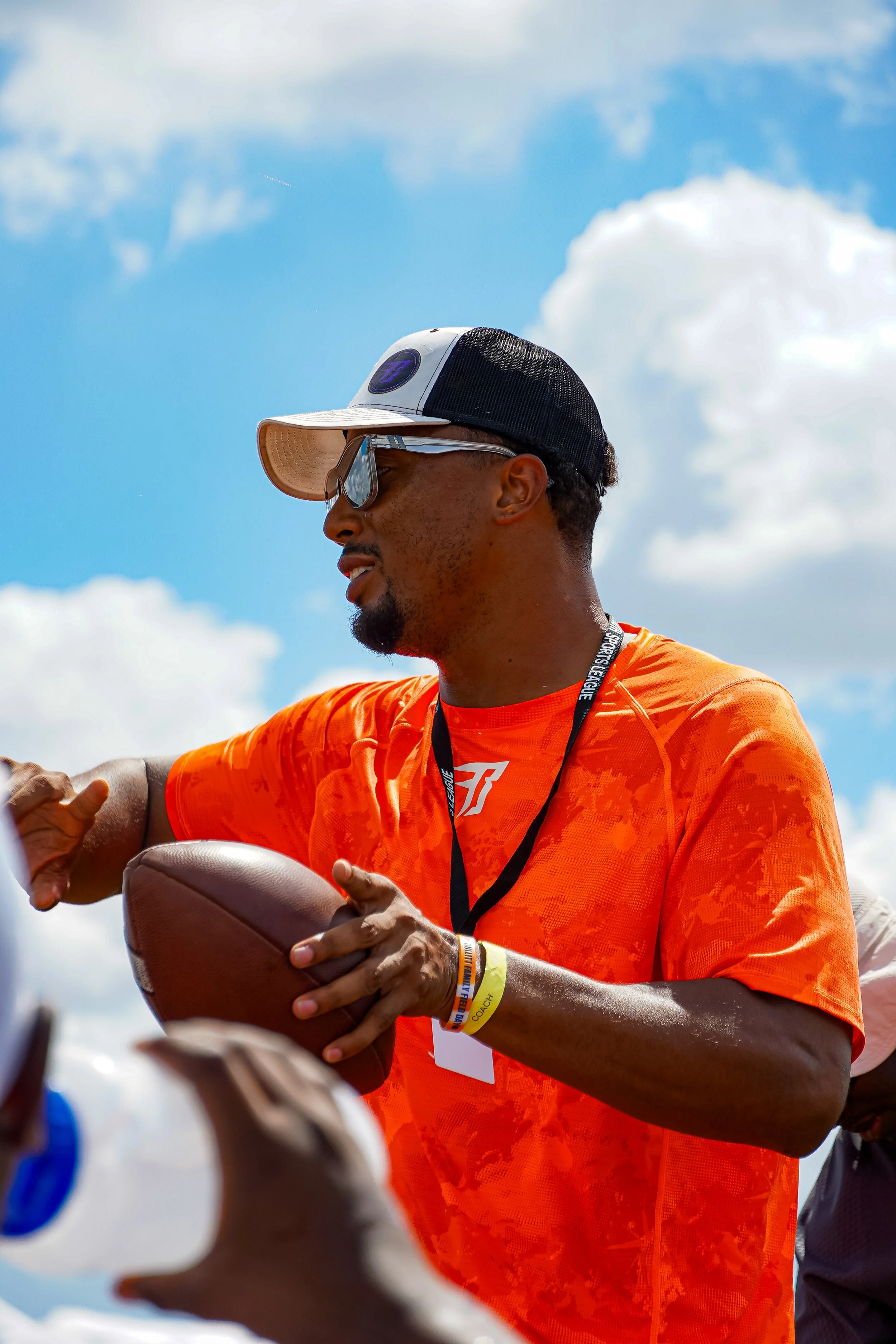 Man in orange shirt holding football while wearing sunglasses, cap, and spectator wristbands outdoors against a blue sky with clouds.