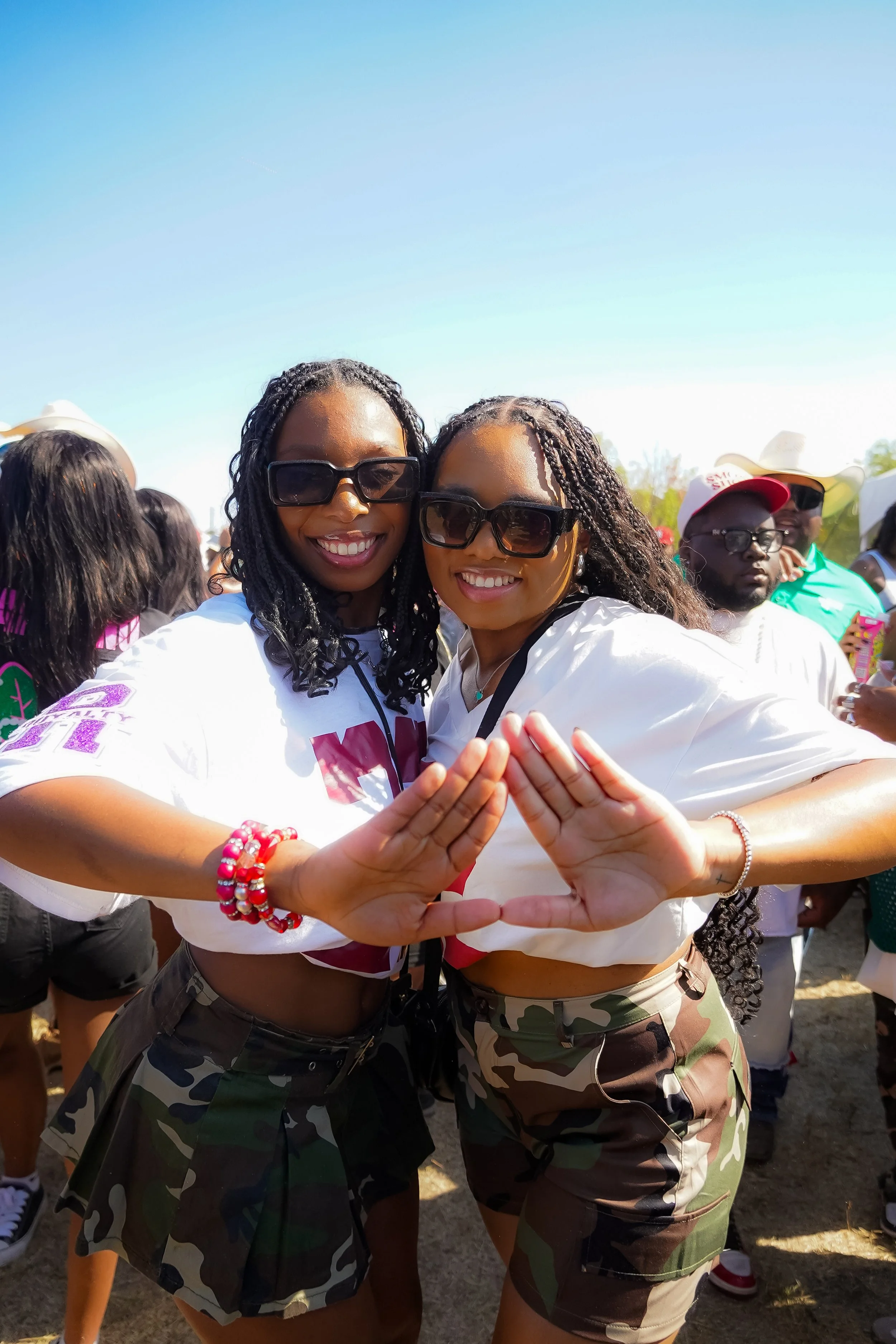 Two smiling women with dark sunglasses, wearing camouflage shorts, are making a triangle shape with their hands together at an outdoor event on a sunny day.