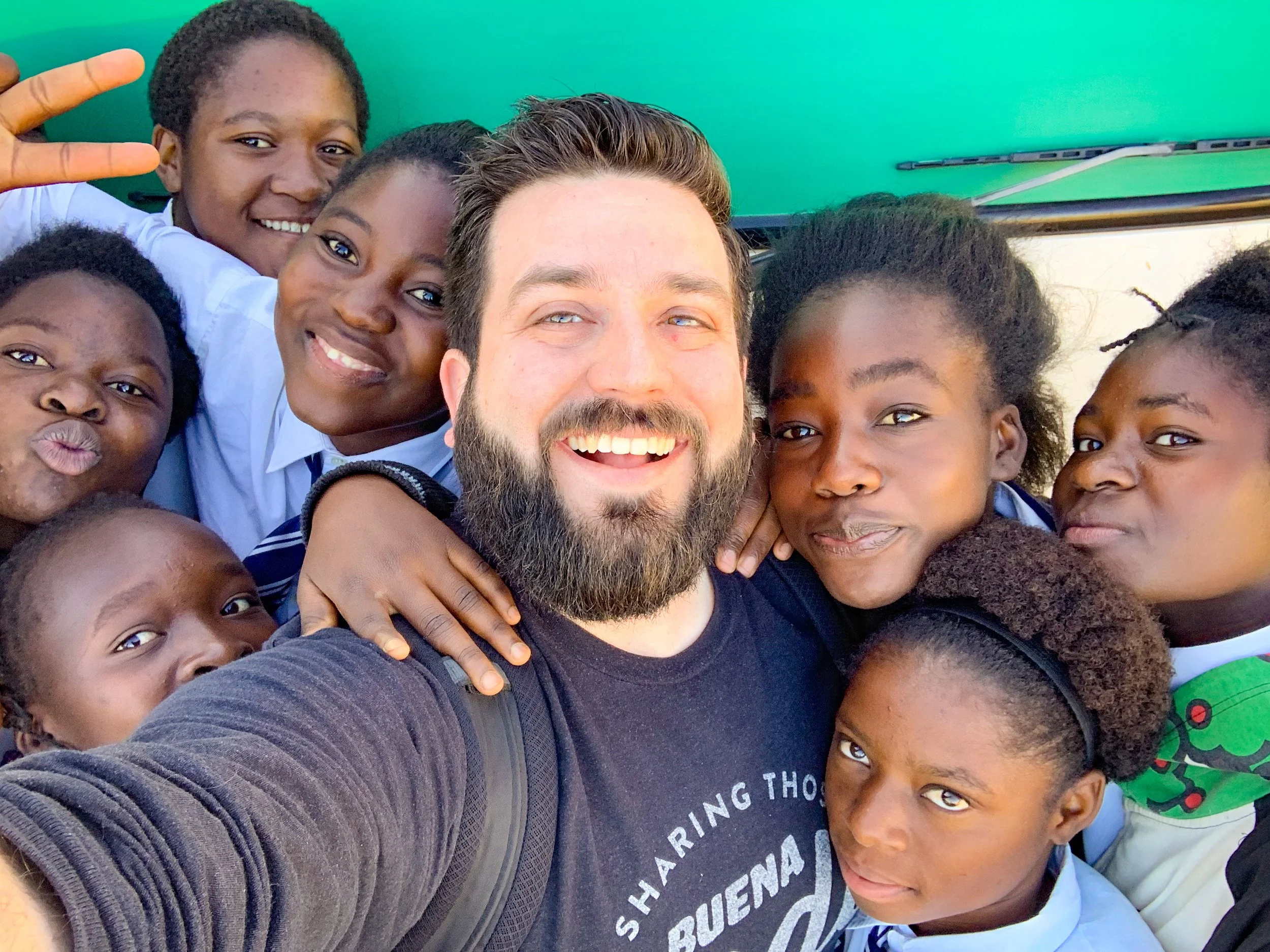 Close-up group selfie of smiling children and an adult man, with some children making peace signs, taken outdoors with a green background.