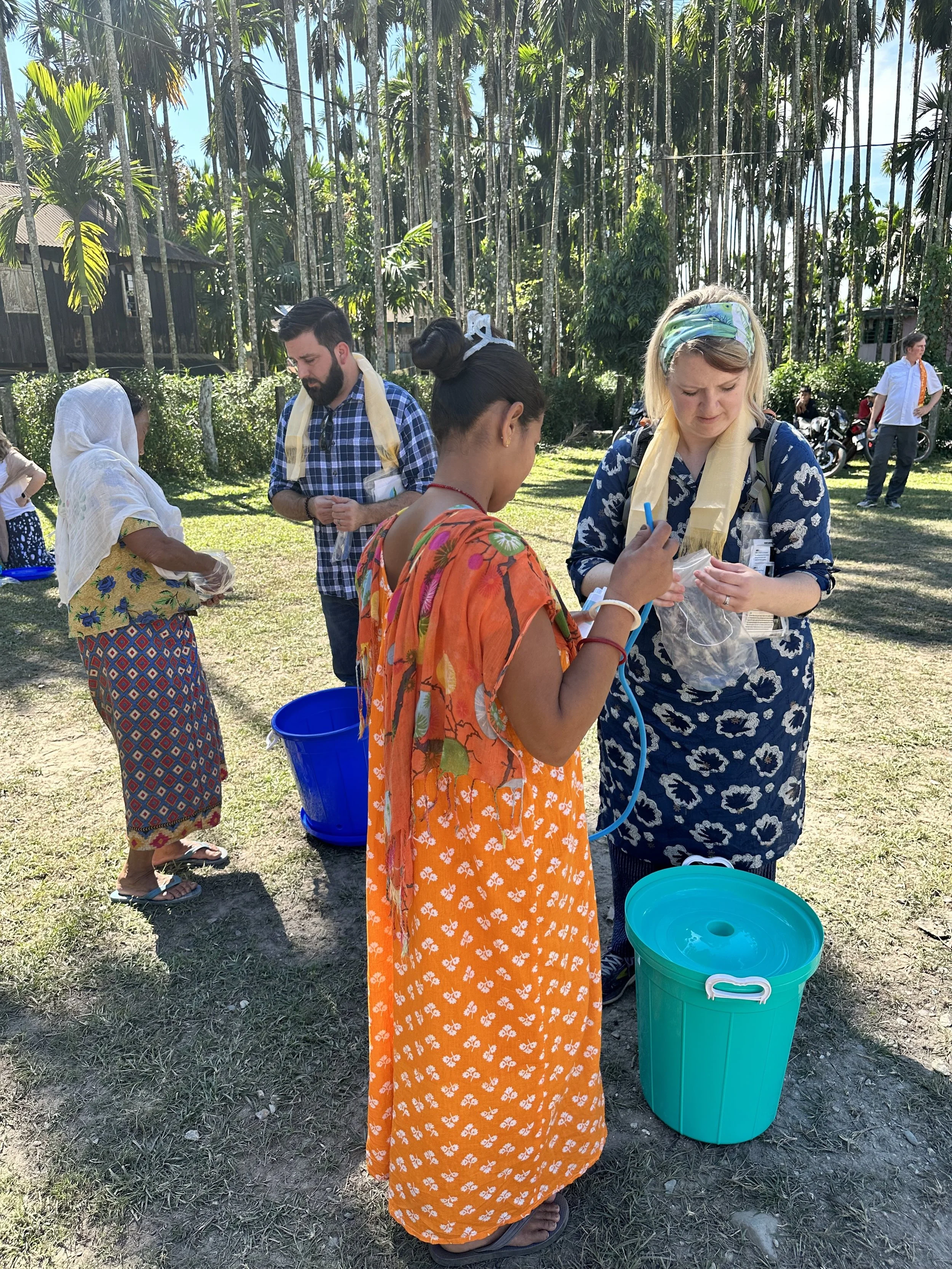 Several people in traditional and casual clothing gather outdoors during the daytime, with tall trees and a building in the background. They appear to be engaging in a community activity involving water containers.