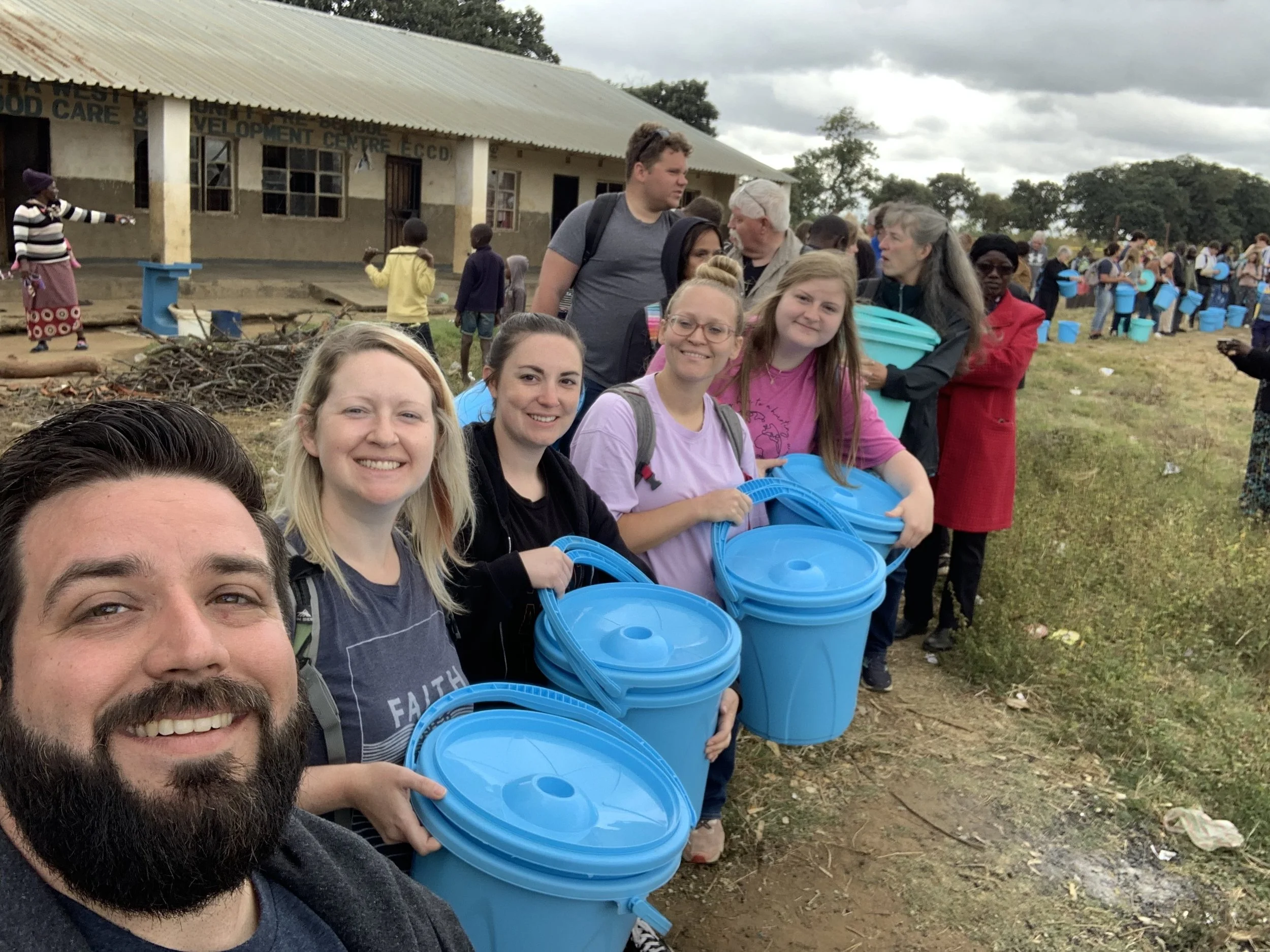 Group of volunteers standing outside with blue buckets, in front of a building with a sign reading 'Development Centre.' Others in the background are also holding blue buckets, and some children are nearby. The sky is overcast.