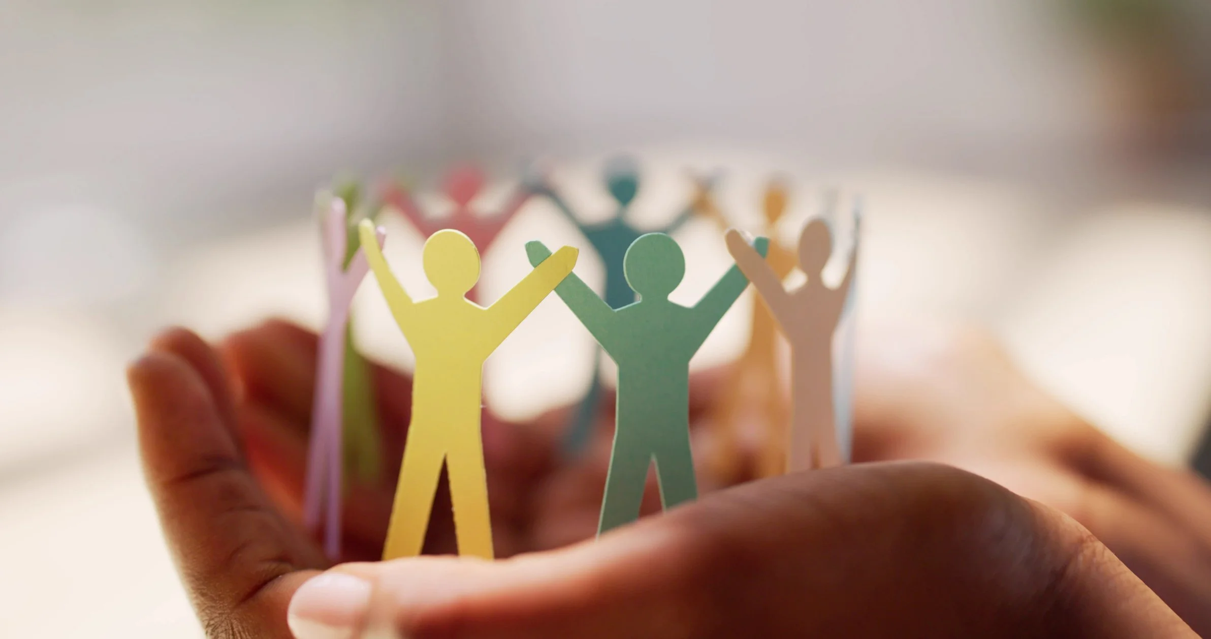 A hand holding cut-out paper figures of people in various colors, forming a circle with arms raised, symbolizing unity and diversity.