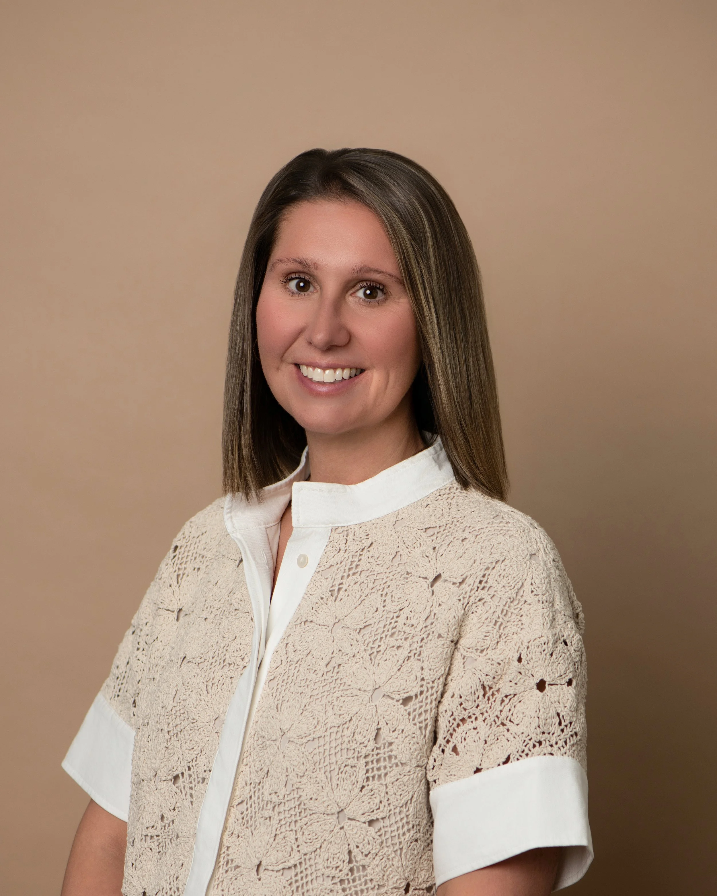 A woman with shoulder-length brown hair, smiling, wearing a white lace top with a high neckline, standing against a plain taupe background.