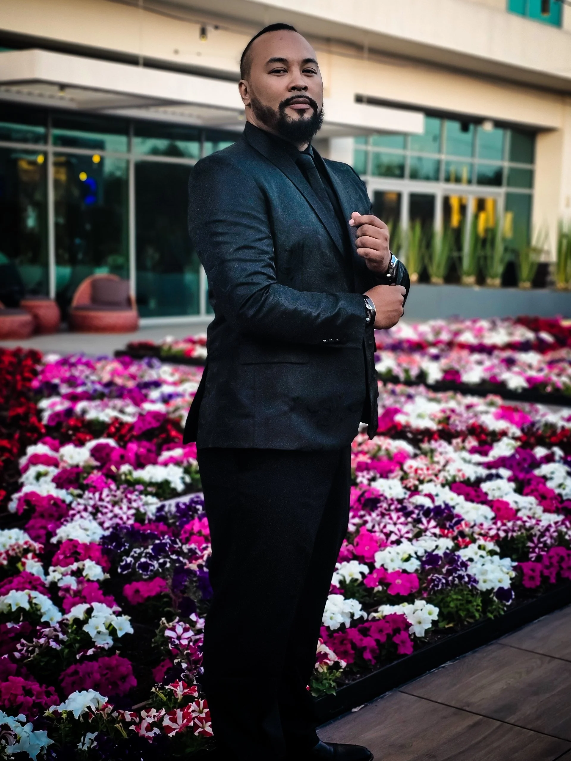 DJ Aeron in a black suit and tie standing outdoors in front of a colorful flower bed with pink, purple, and white flowers, with a building and large glass windows in the background.