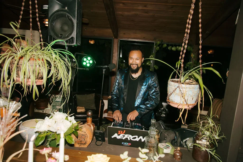 DJ Aeron smiling at a setup of DJ equipment, surrounded by hanging and potted plants, in a cozy indoor space.