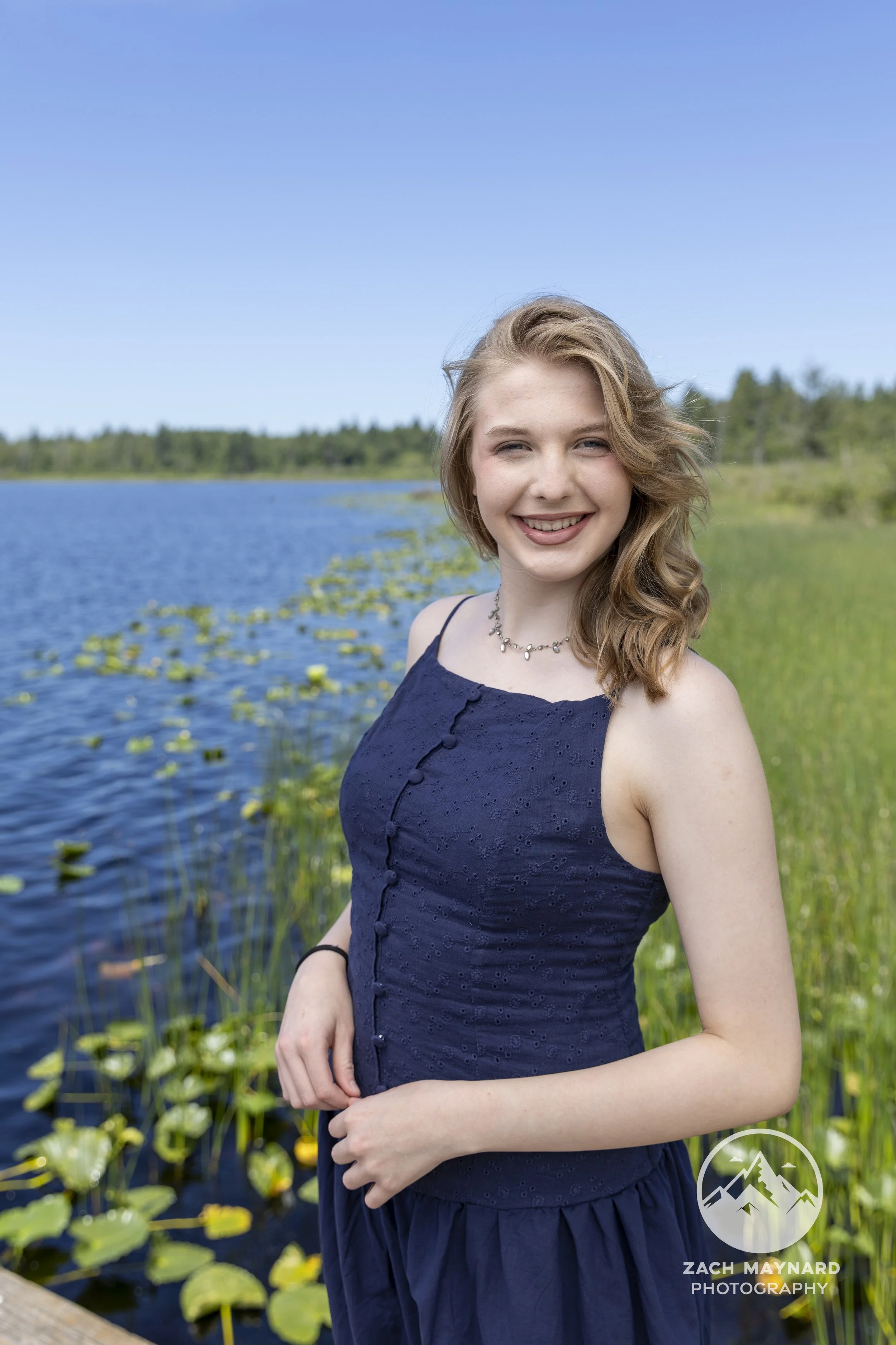 Smiling teen girl in a navy dress by a lily-pad lake during a summer senior portrait session in Snohomish County