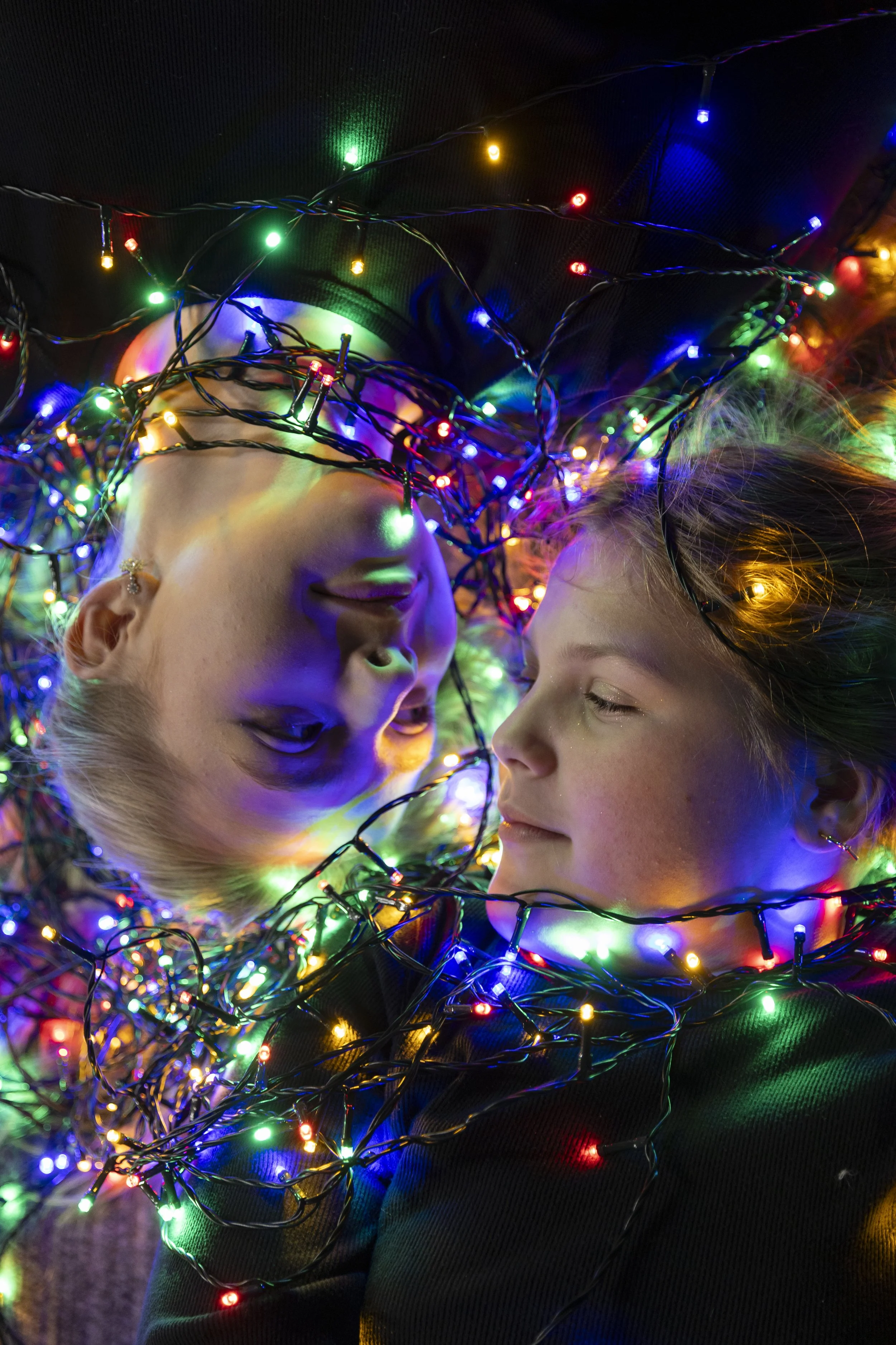 Creative holiday portrait of two children wrapped in colorful Christmas lights, featuring vibrant festive lighting and candid family photography.