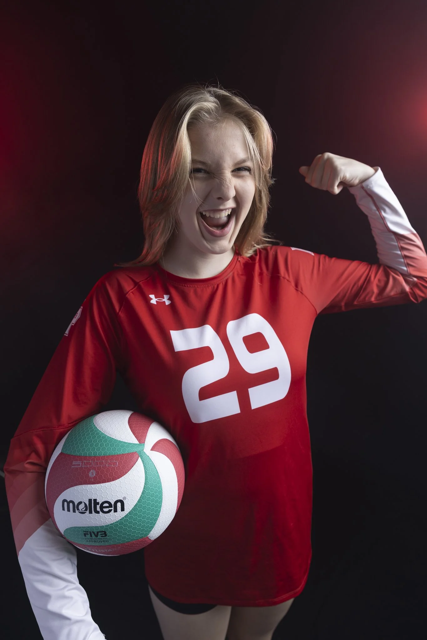 A young woman in a red volleyball uniform flexes her right arm and holds a colorful volleyball in her left hand, standing against a black background.