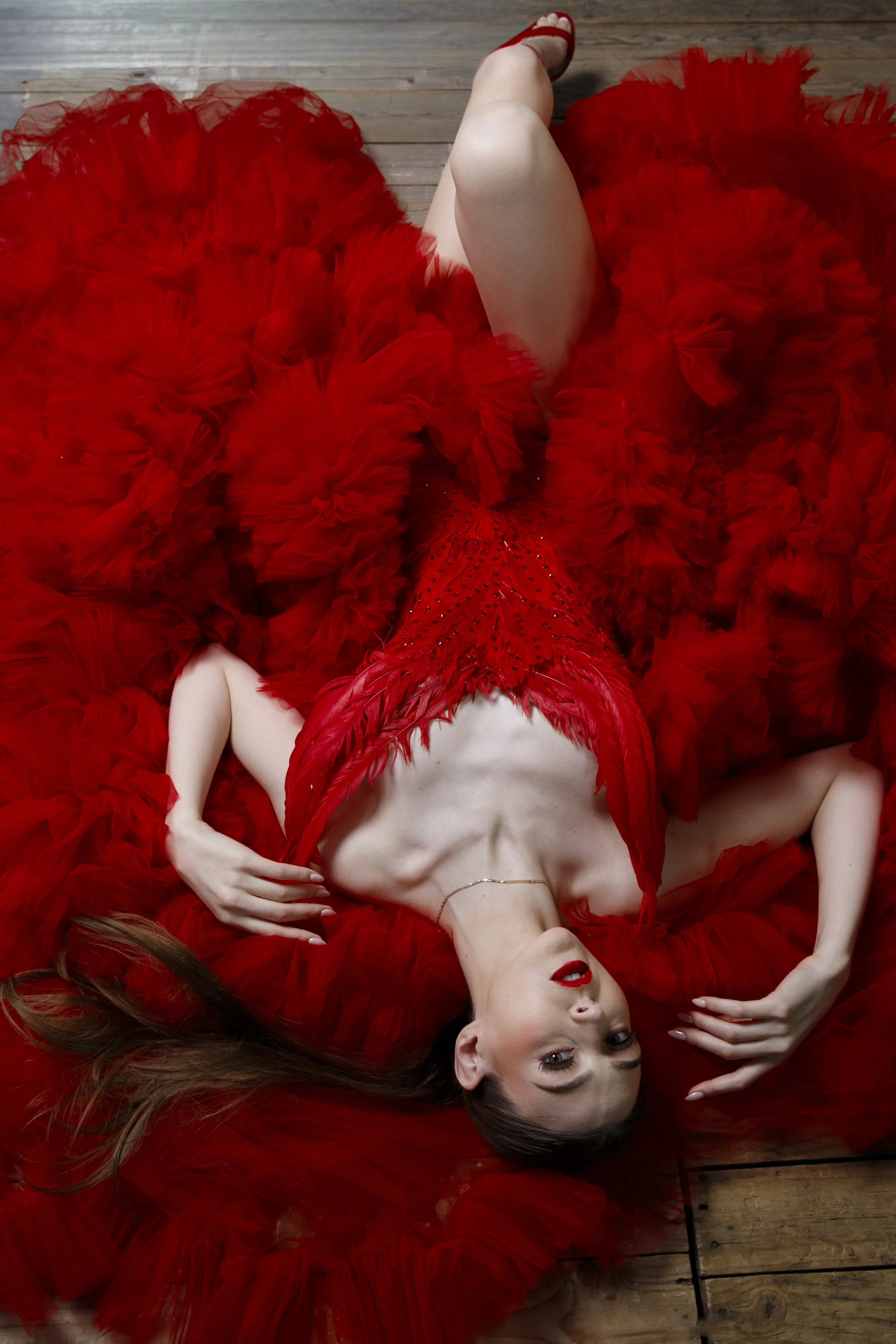 A woman lying on a wooden floor, dressed in a red feathered gown with ruffles, with her head tilted and one arm resting on her stomach.