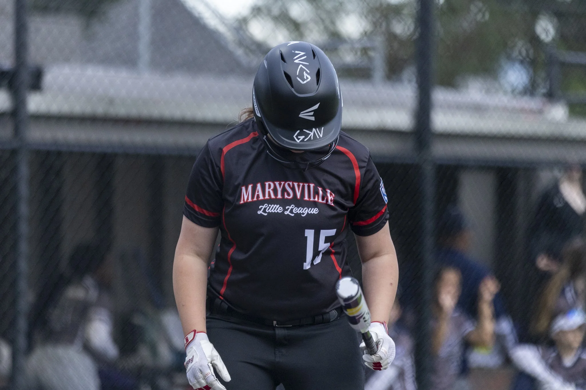 A female baseball player wearing a black helmet and a black jersey with red accents and the text 'MARYSVILLE Little League' on it, standing on the field with a bat.