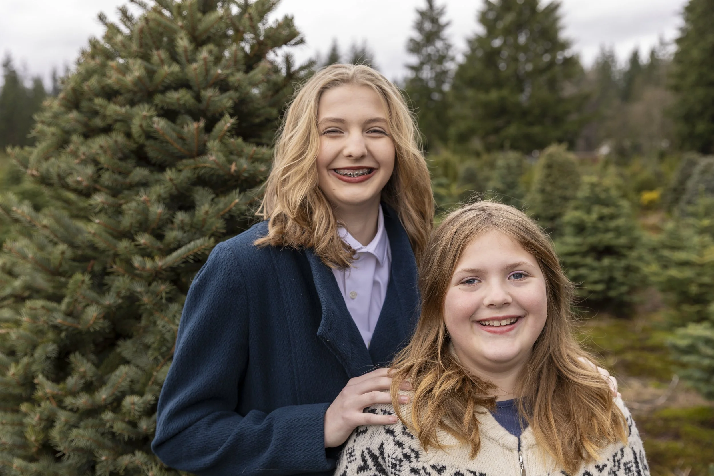 Two young girls with blonde hair smiling in a Christmas tree farm, surrounded by evergreen trees.