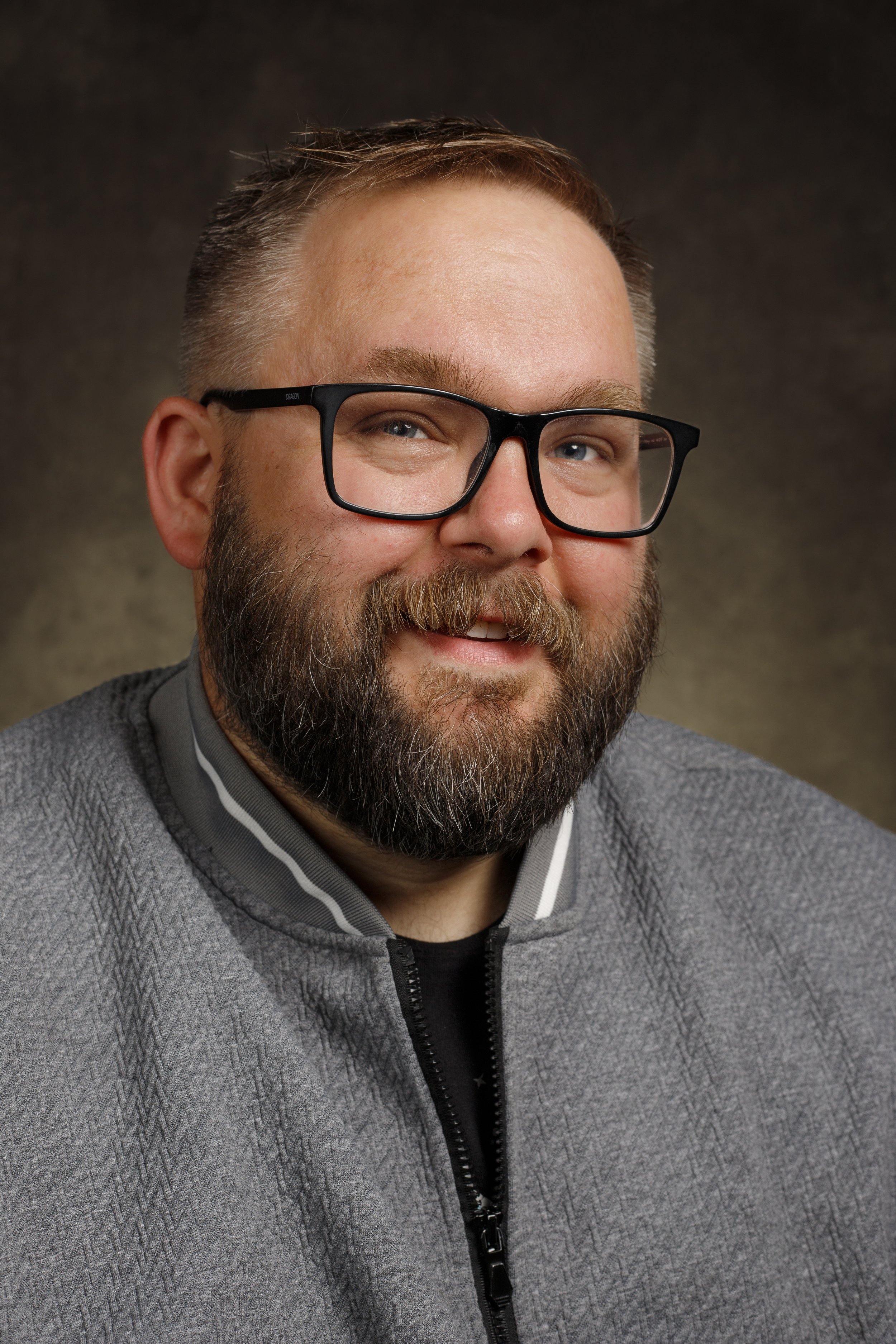 Portrait of a man with glasses, a beard, and a short hairstyle, wearing a gray jacket with a black shirt, smiling against a dark, textured background.