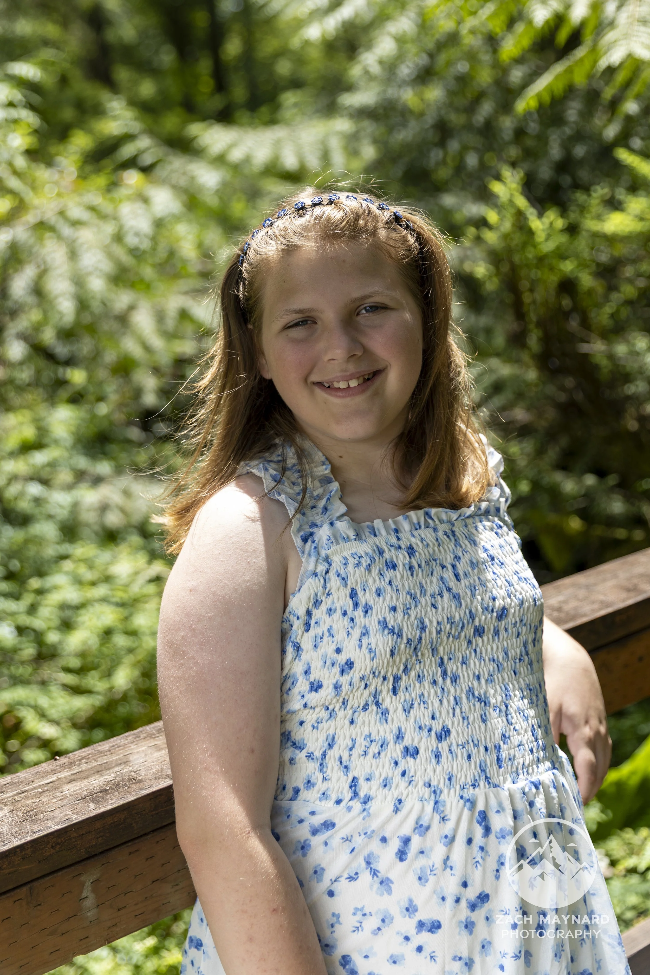 Preteen girl in a blue floral dress smiling on a wooden bridge in a forest during a summer portrait session in Snohomish County